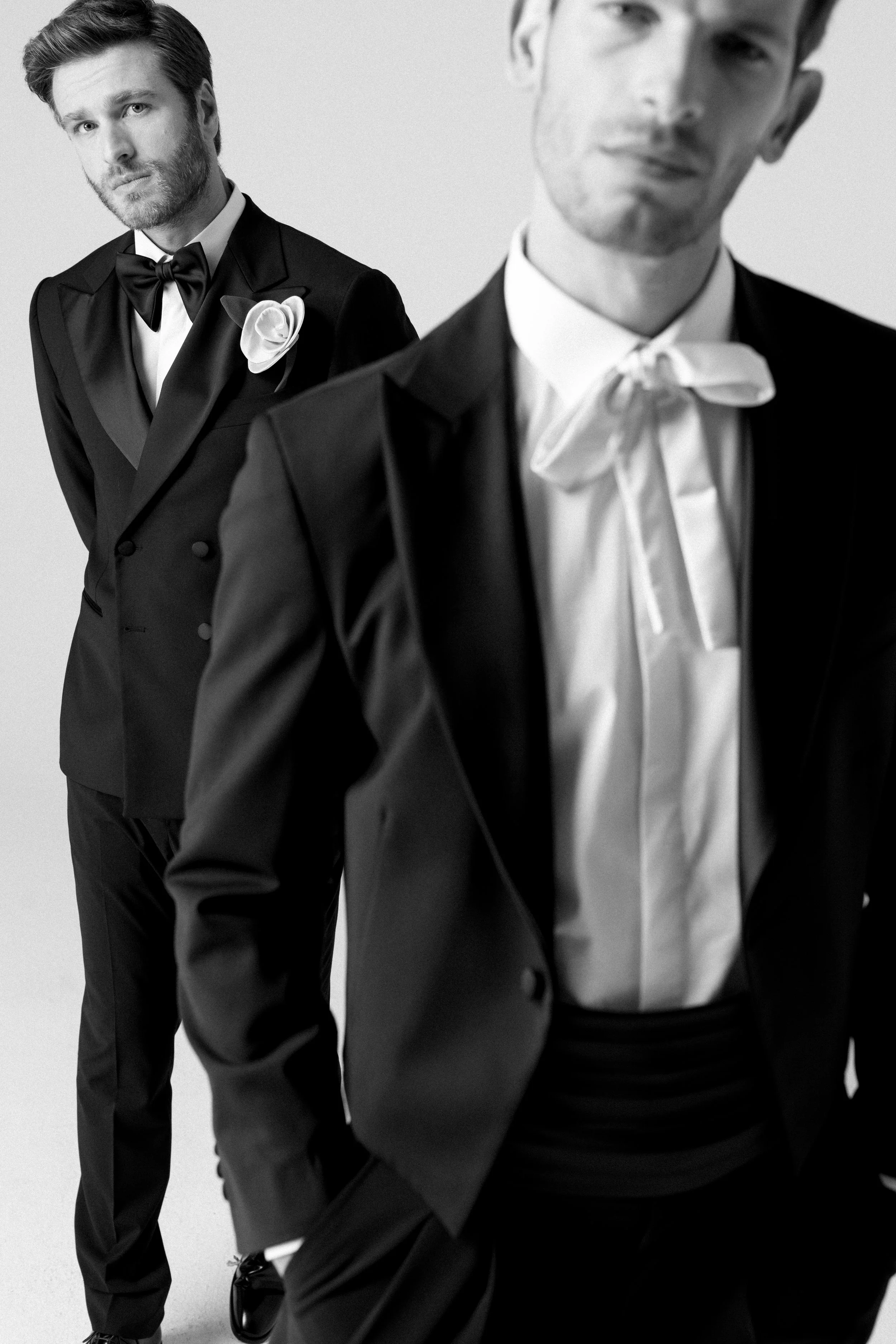 Due uomini vestiti in smoking eleganti, uno con fiocco nero e l'altro con cravatta bianca, in uno scatto in bianco e nero, con uno sfondo chiaro.