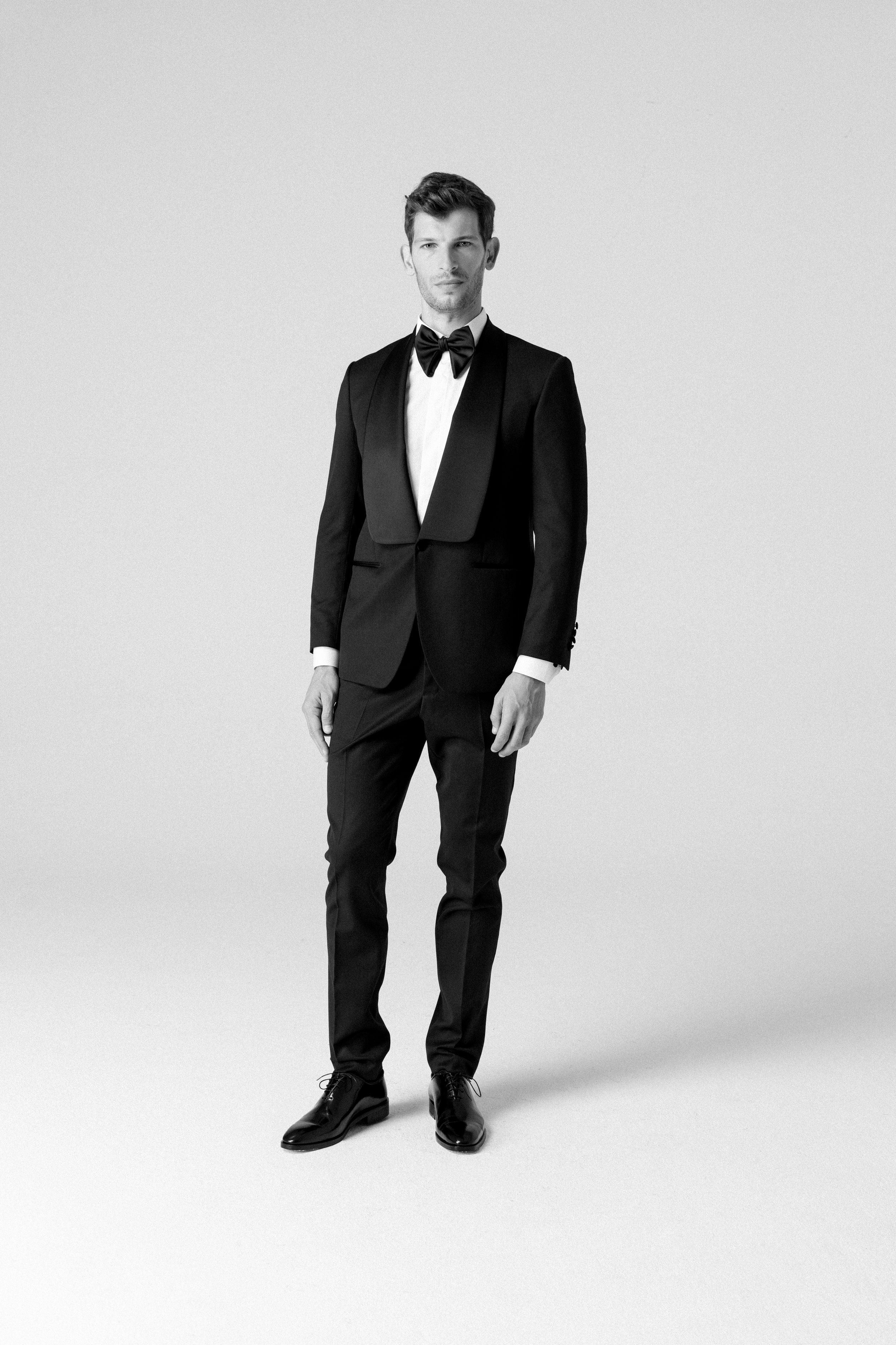 Uomo con abito da sera nero con papillon, in piedi su sfondo chiaro, ritratto in bianco e nero.
