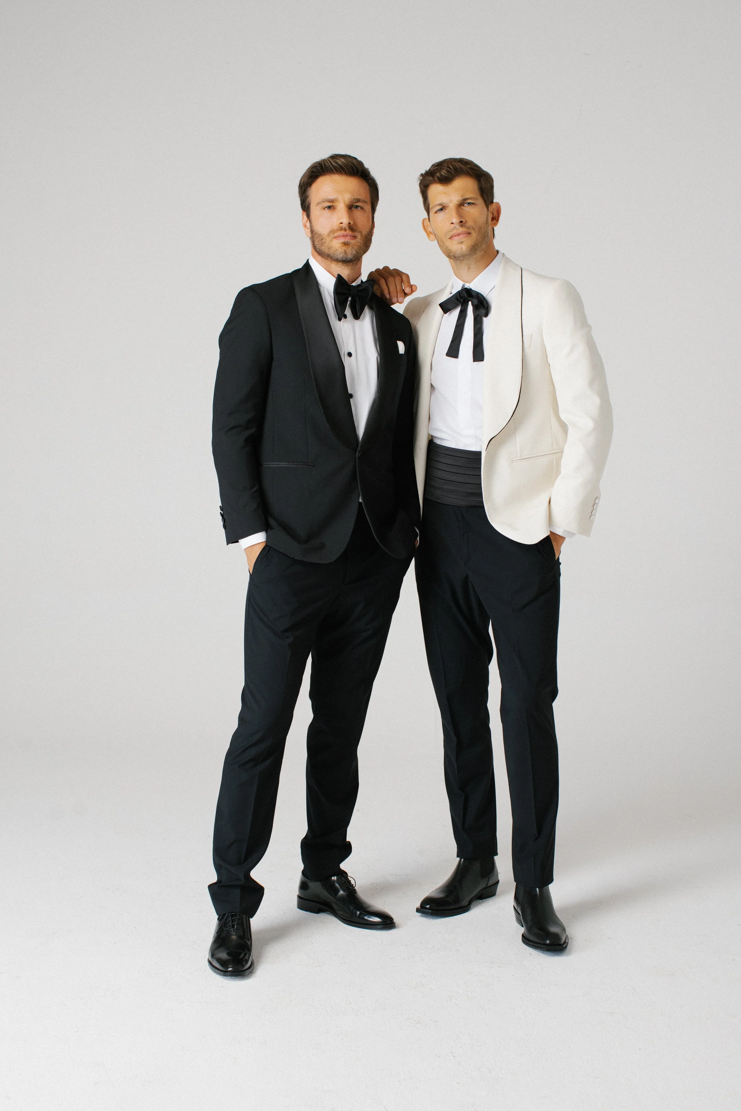 Due uomini eleganti con abiti da sera formali, uno in completo nero con bottoni e l'altro in completo bianco con pantaloni neri, entrambi indossano papillon nere e stanno in piedi su uno sfondo grigio chiaro.