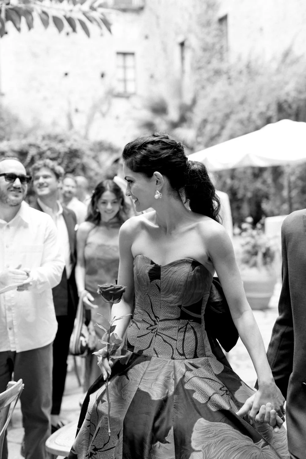 Donna sorridente con abito floreale in un evento all'aperto con persone intorno, in bianco e nero.