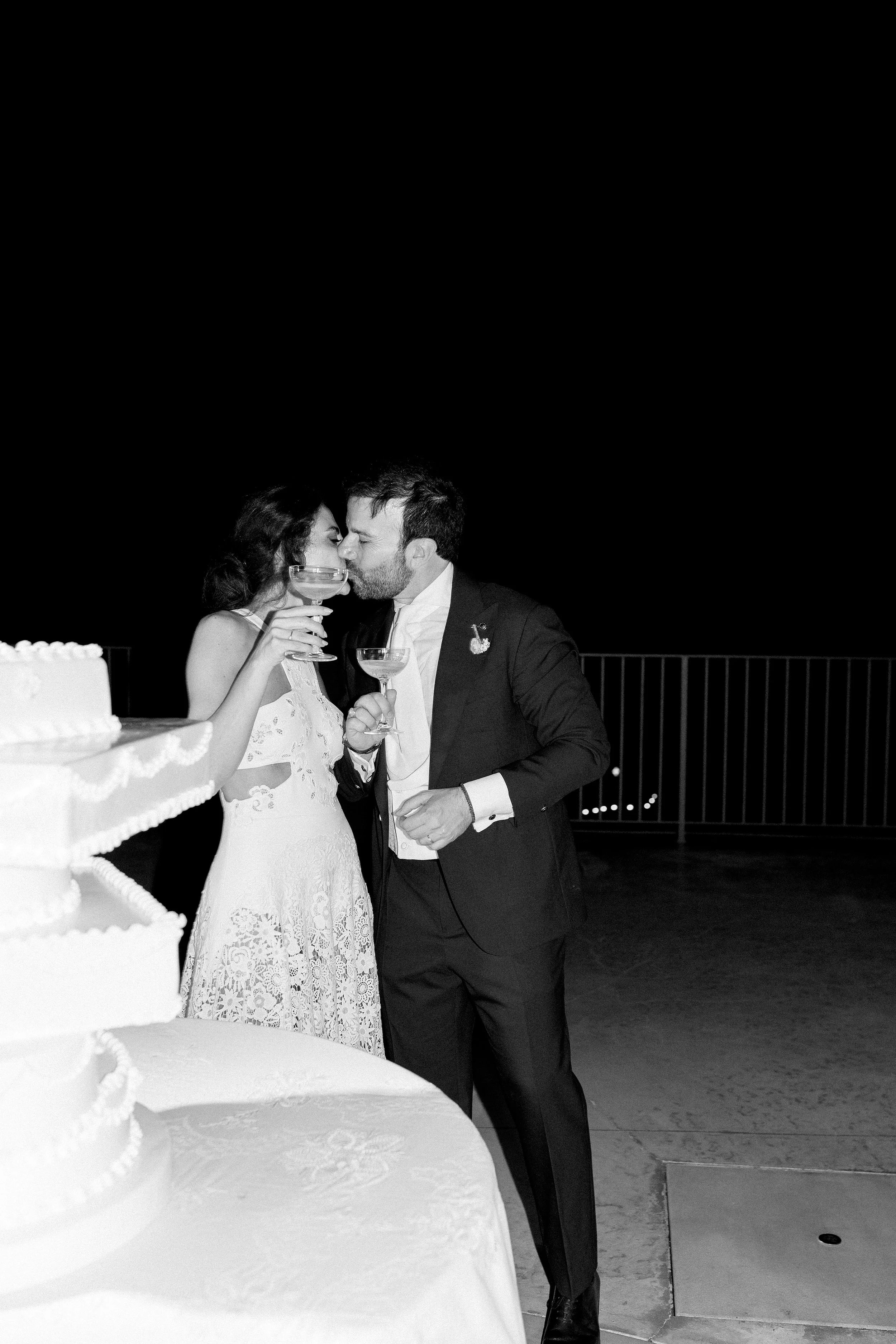 Una coppia di sposi si bacia durante il loro matrimonio, con una torta nuziale visibile in primo piano.
