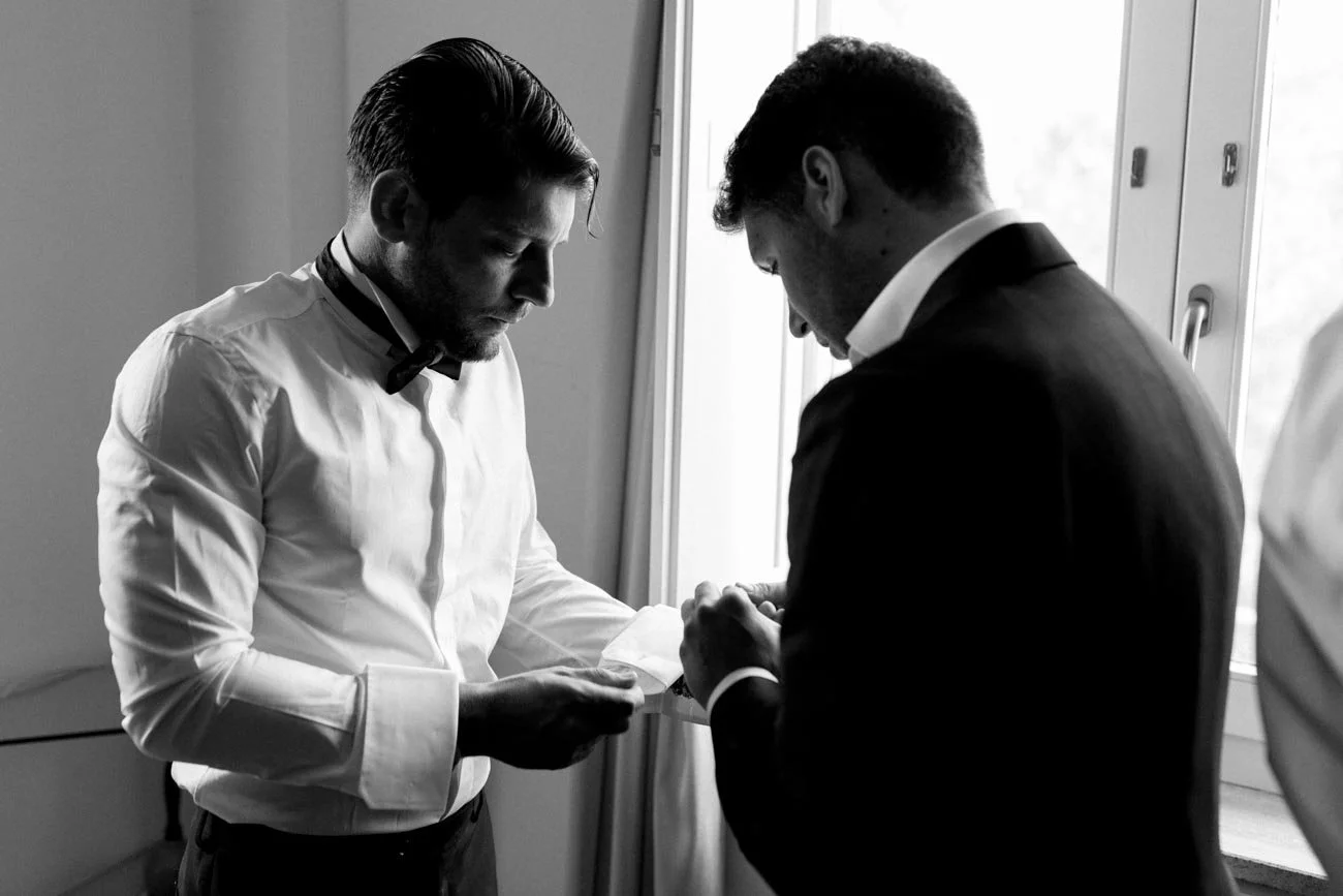 Due uomini eleganti, uno con camicia bianca e l'altro con giacca nera, si scambiano promesse o anelli in una stanza con finestra