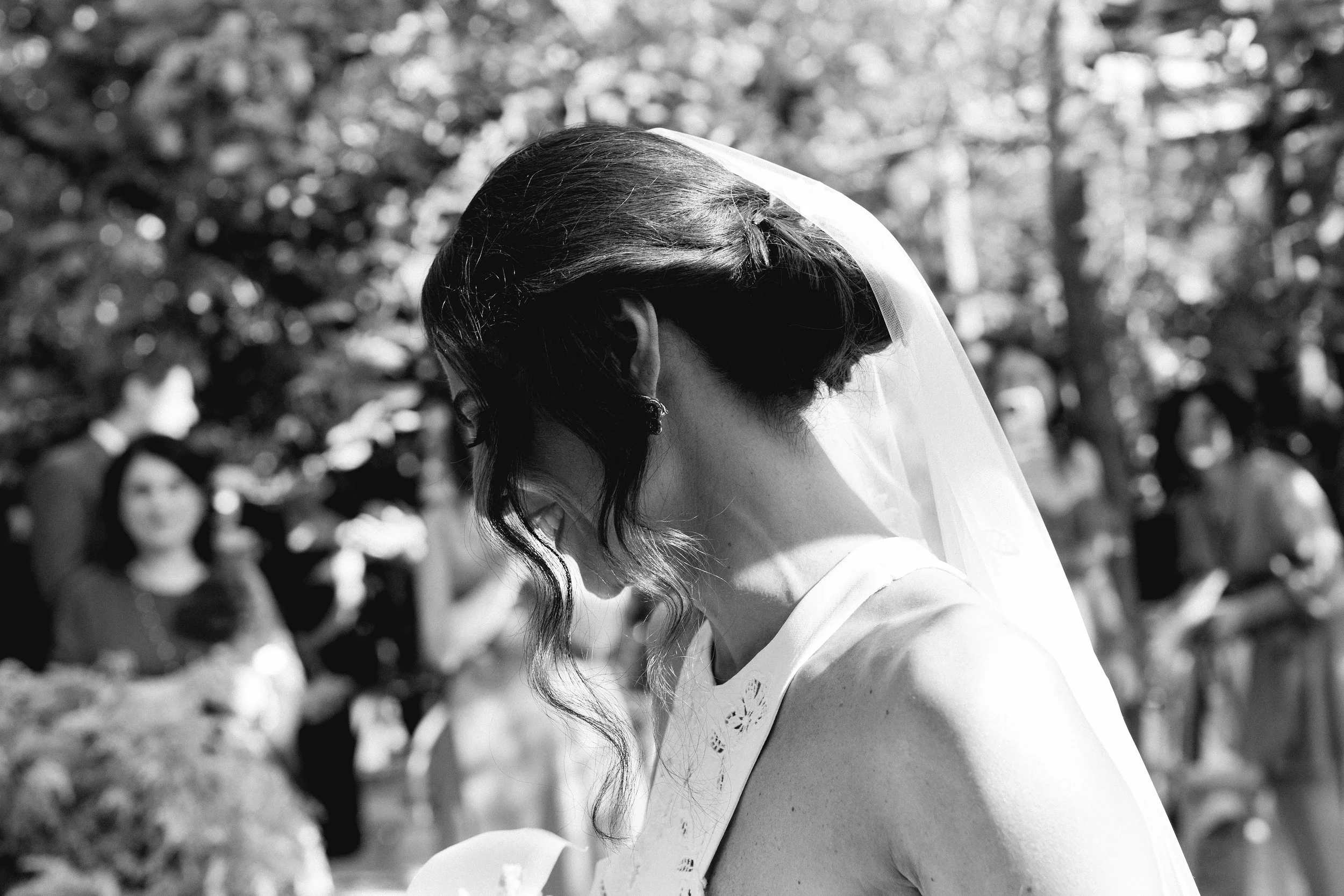 Una donna con un velo da sposa in un matrimonio all'aperto, vista di profilo, con amici in sottofondo.