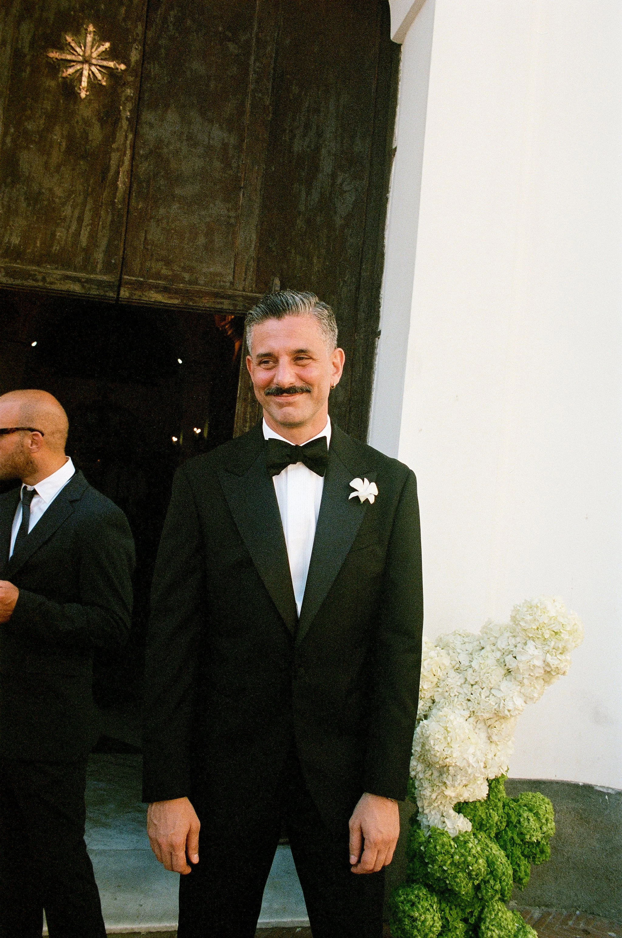 Uomo in completo nero con farfallino nero e fiore bianco sulla giacca, sorridente, in un evento formale vicino a decorazioni di fiori bianchi e verdi.