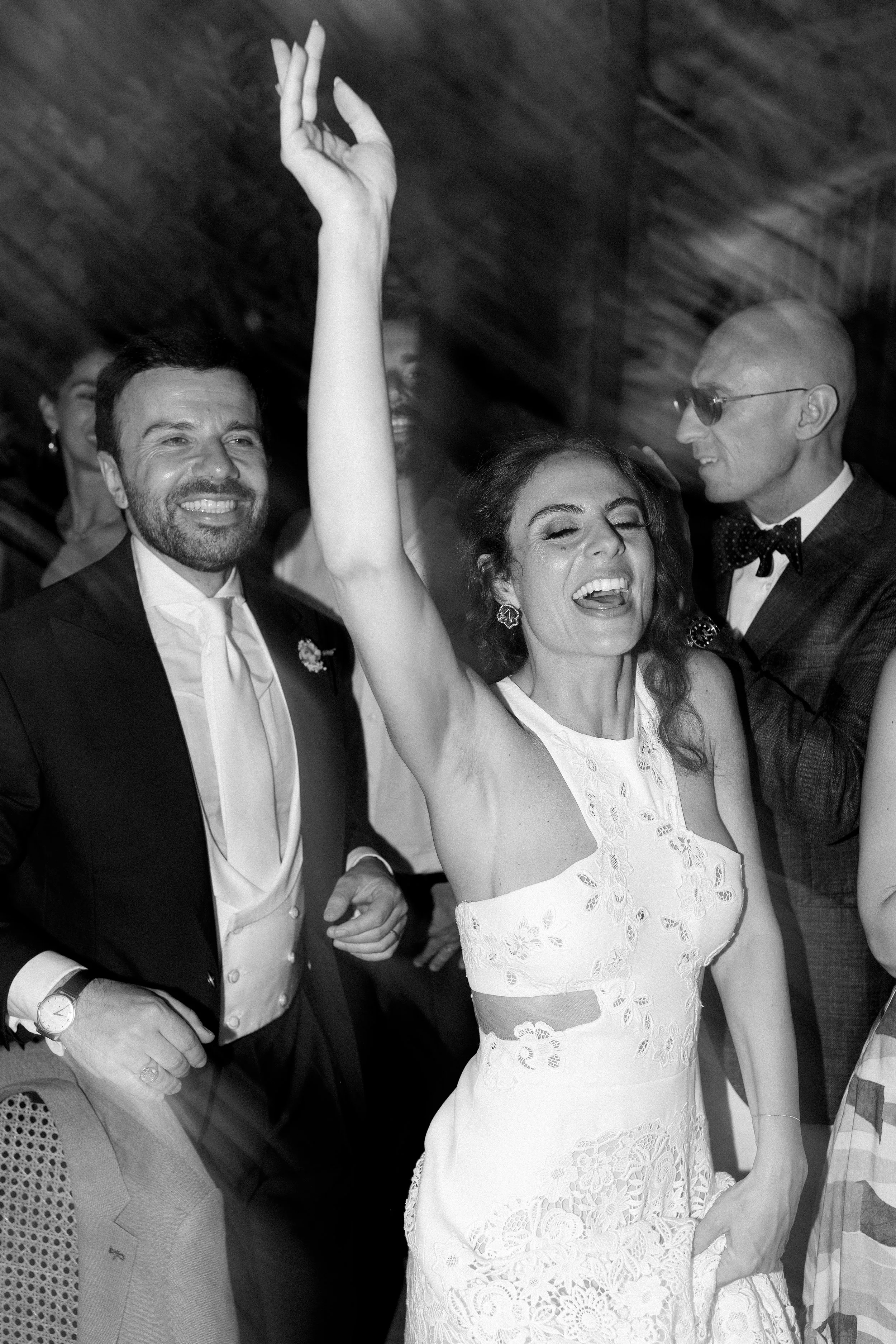 Persone che festeggiano, donna che balla e sorride in primo piano, uomini eleganti sullo sfondo in un evento sociale.