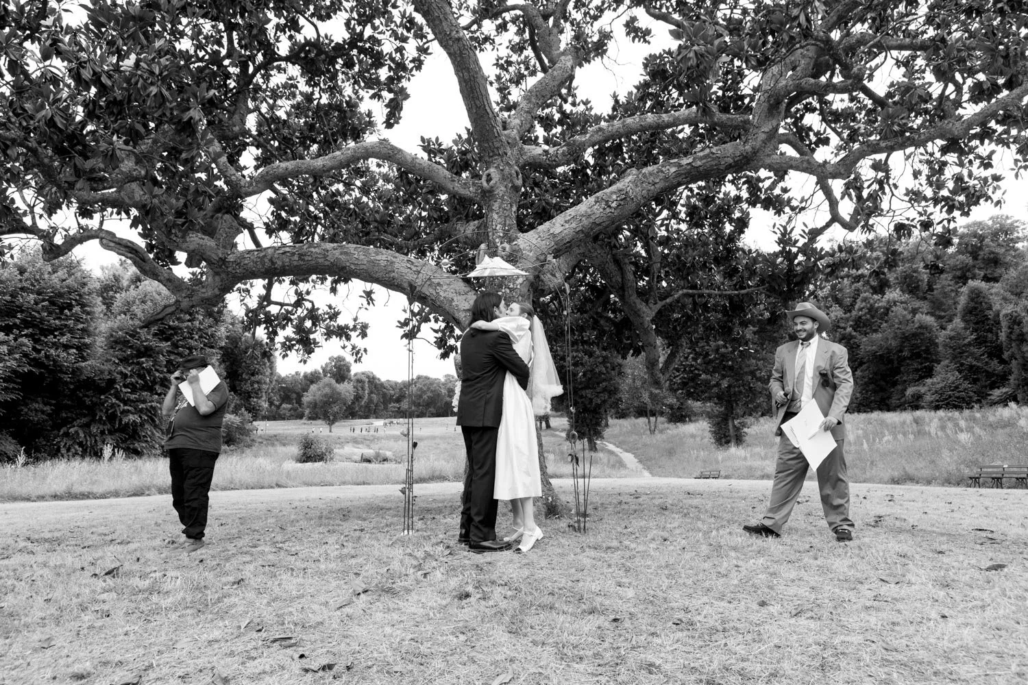 Un matrimonio si bacia sotto un grande albero in un parco, con un officiatore a sinistra che legge un documento e un uomo con cappello a destra che sorride.