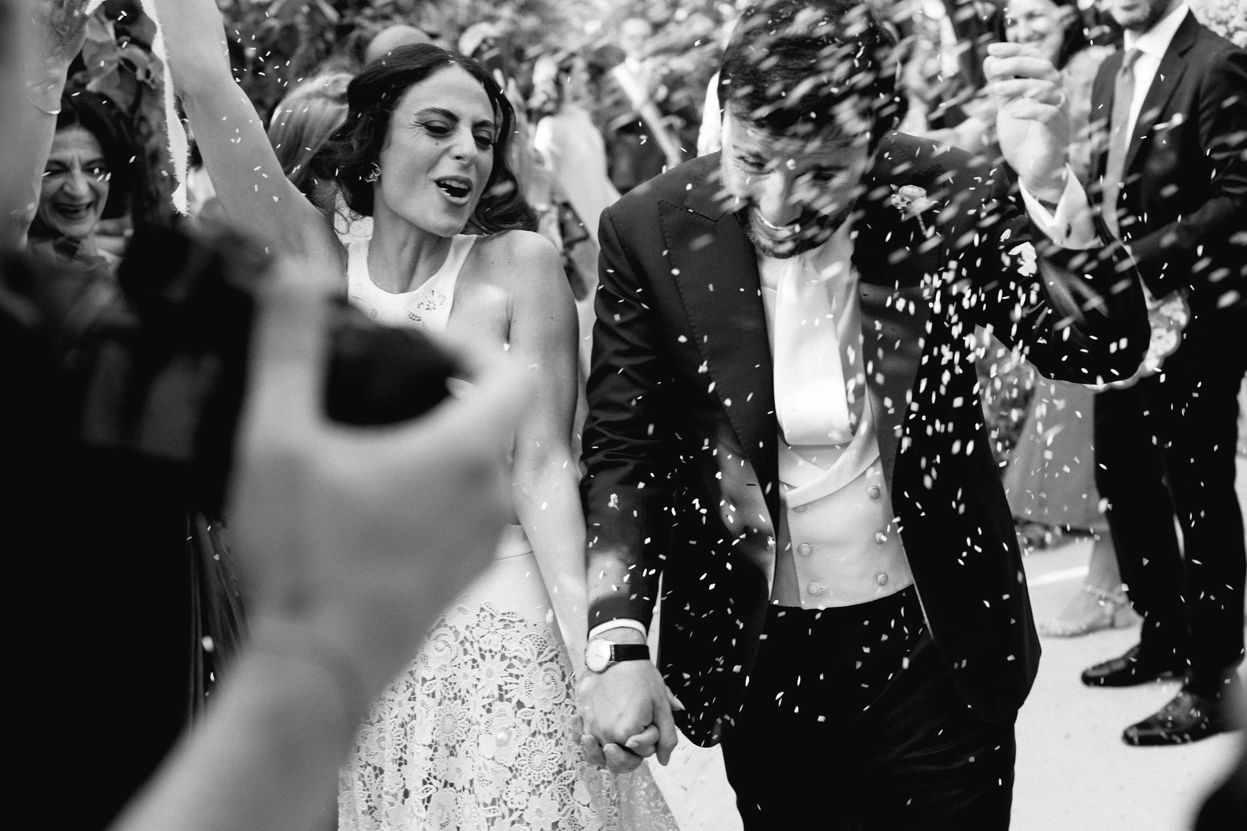 Una coppia di sposi sorride mentre viene festeggiata dai partecipanti, con coriandoli che cadono.