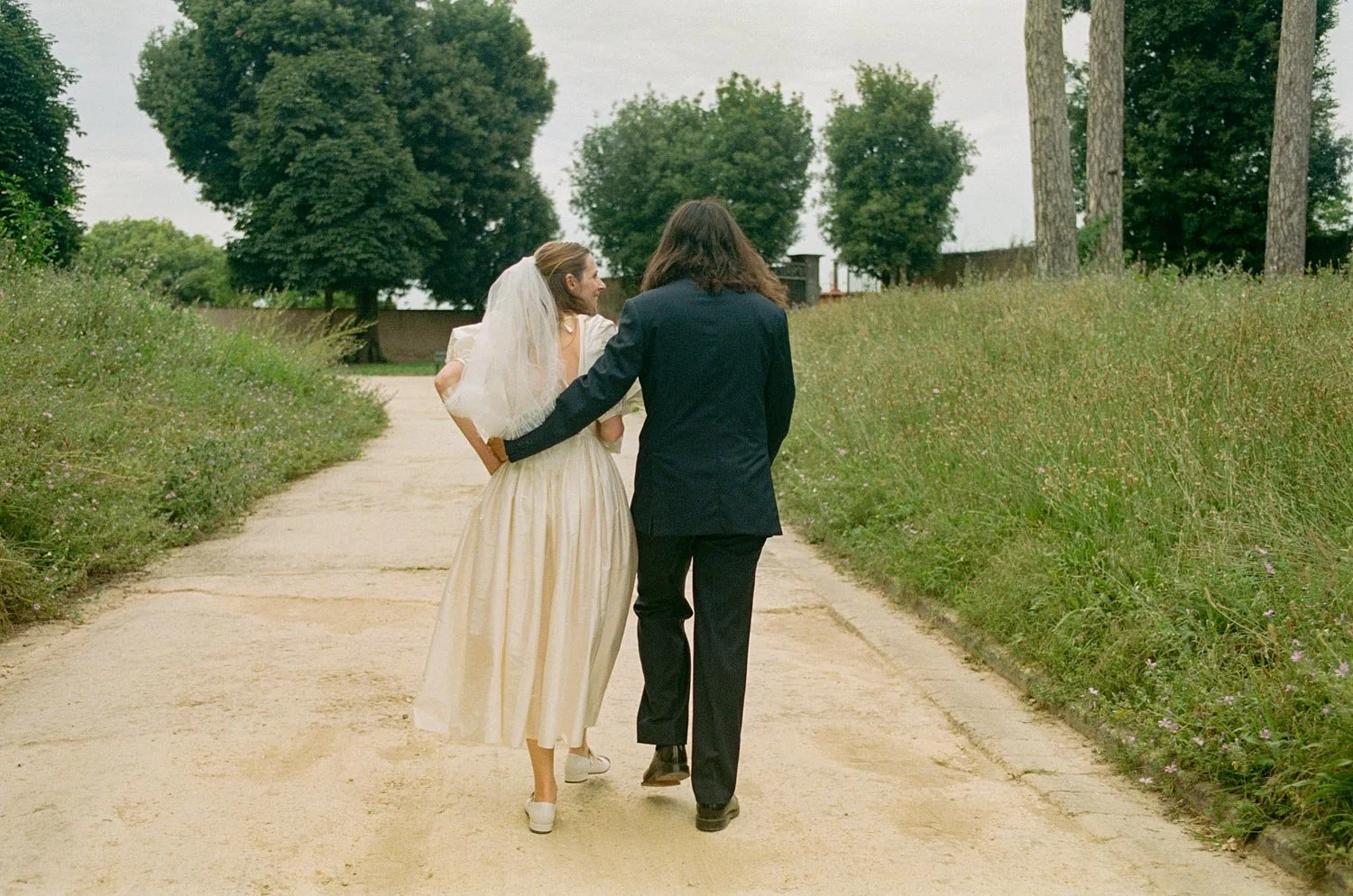 Coppia di sposi che cammina lungo un sentiero in un parco, con il braccio attorno alle spalle l'uno dell'altra, immersi in una giornata nuvolosa.