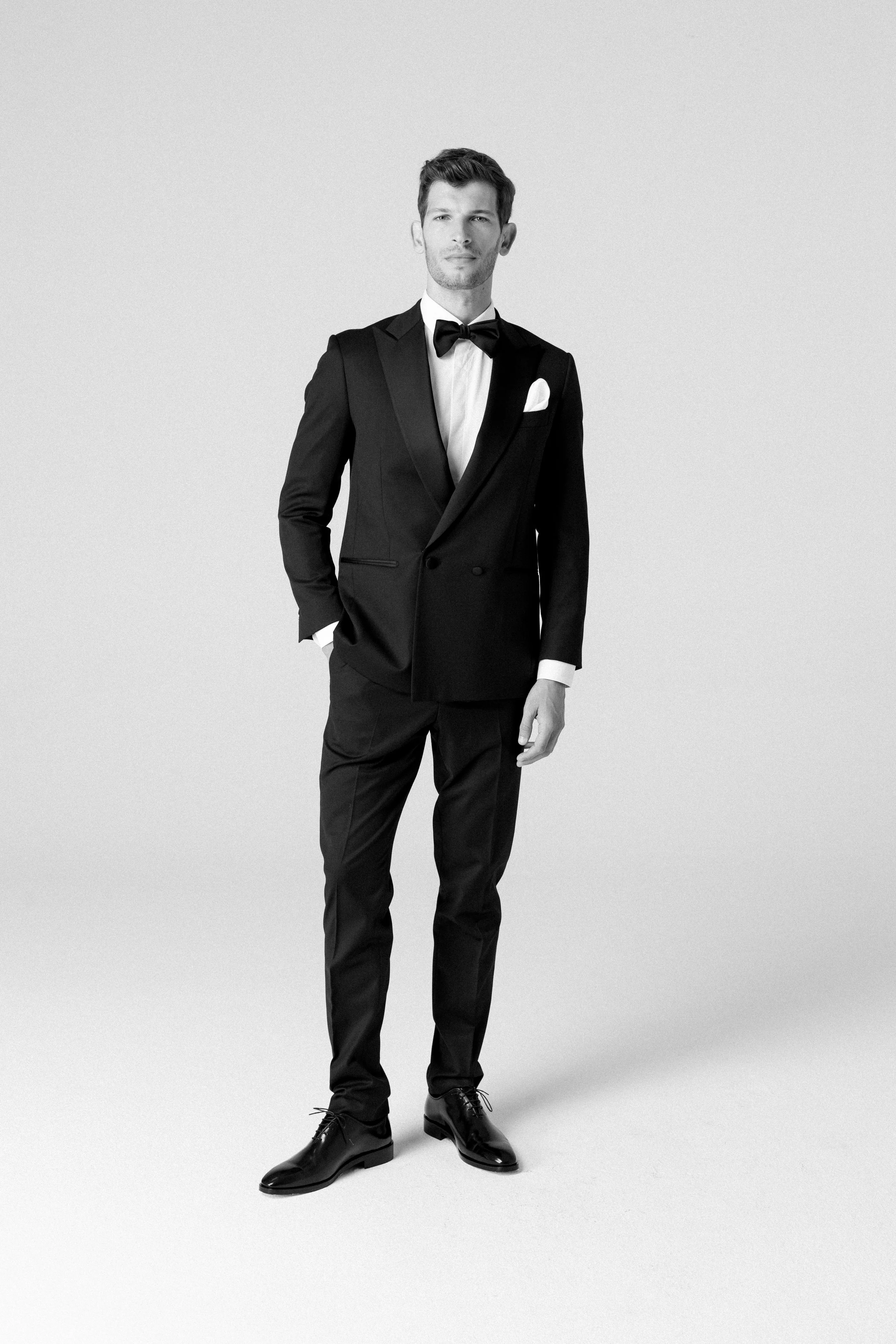 Uomo in abito da sera nero con camicia bianca, papillon nero e pochette bianca, in piedi su sfondo neutro.