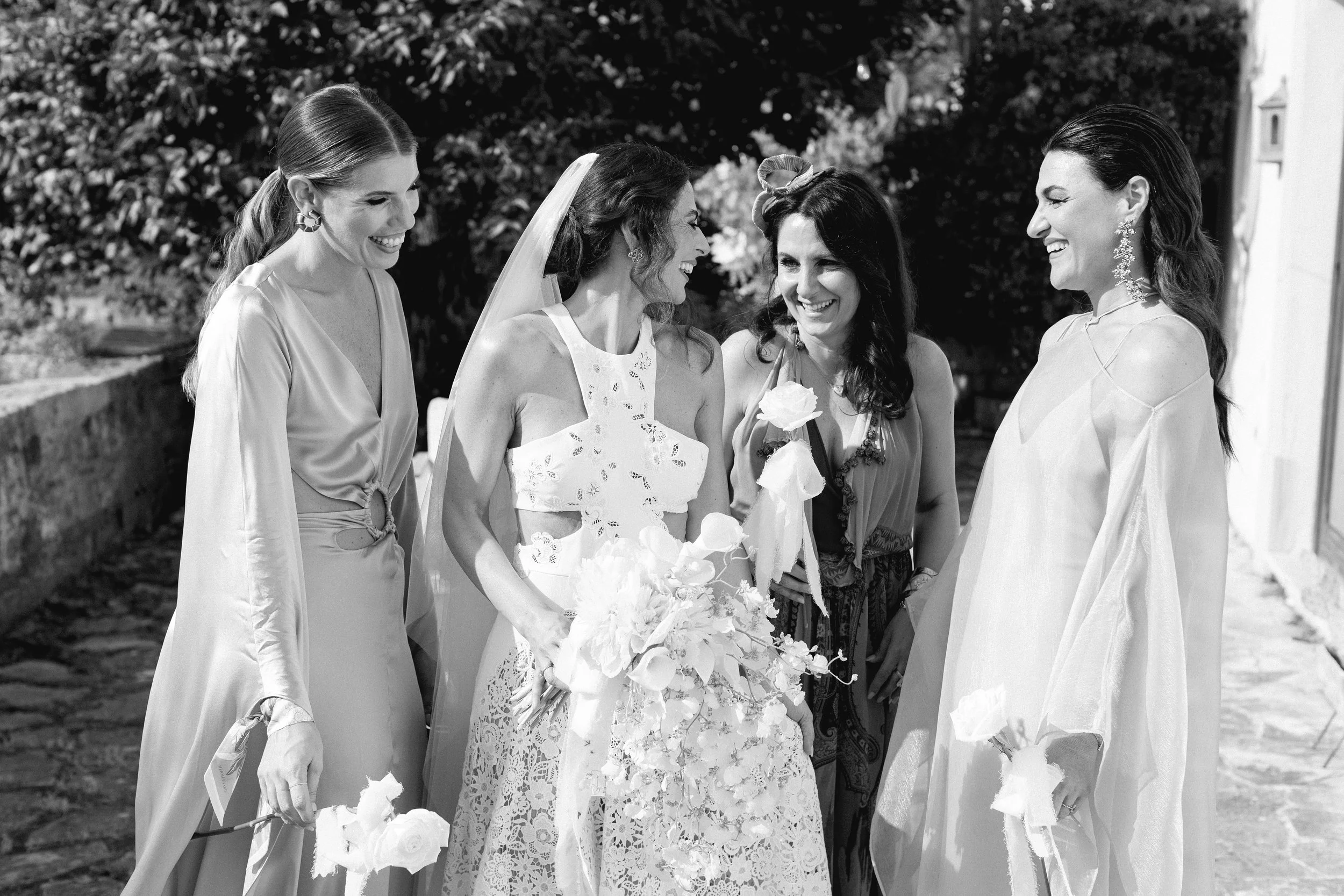 Gruppo di cinque donne sorridenti in abiti eleganti, con una in abito da sposa, che si scambiano sorrisi e fiori all'aperto