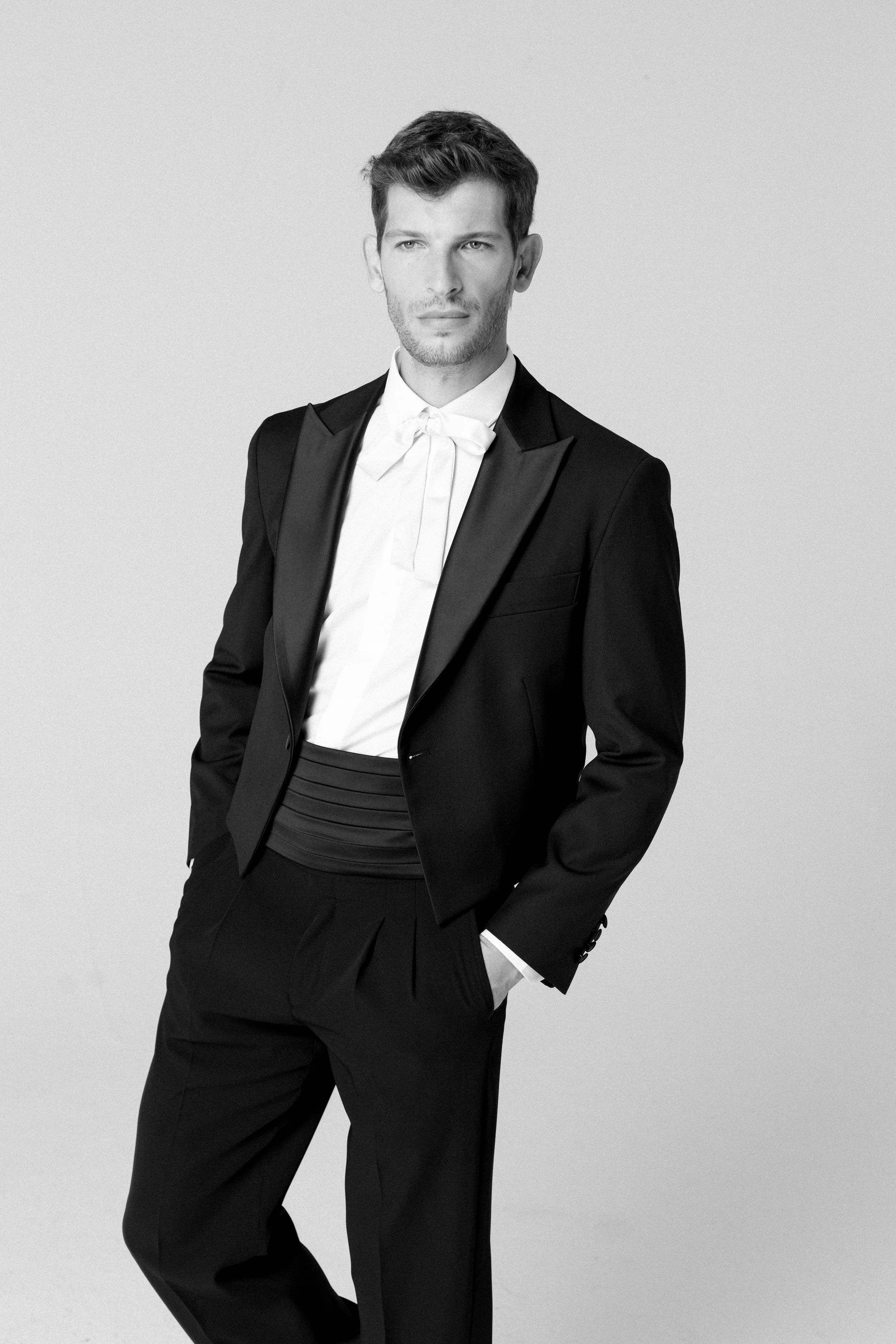Uomo in abito nero con cravatta bianca, posa con una mano in tasca davanti a uno sfondo neutro.