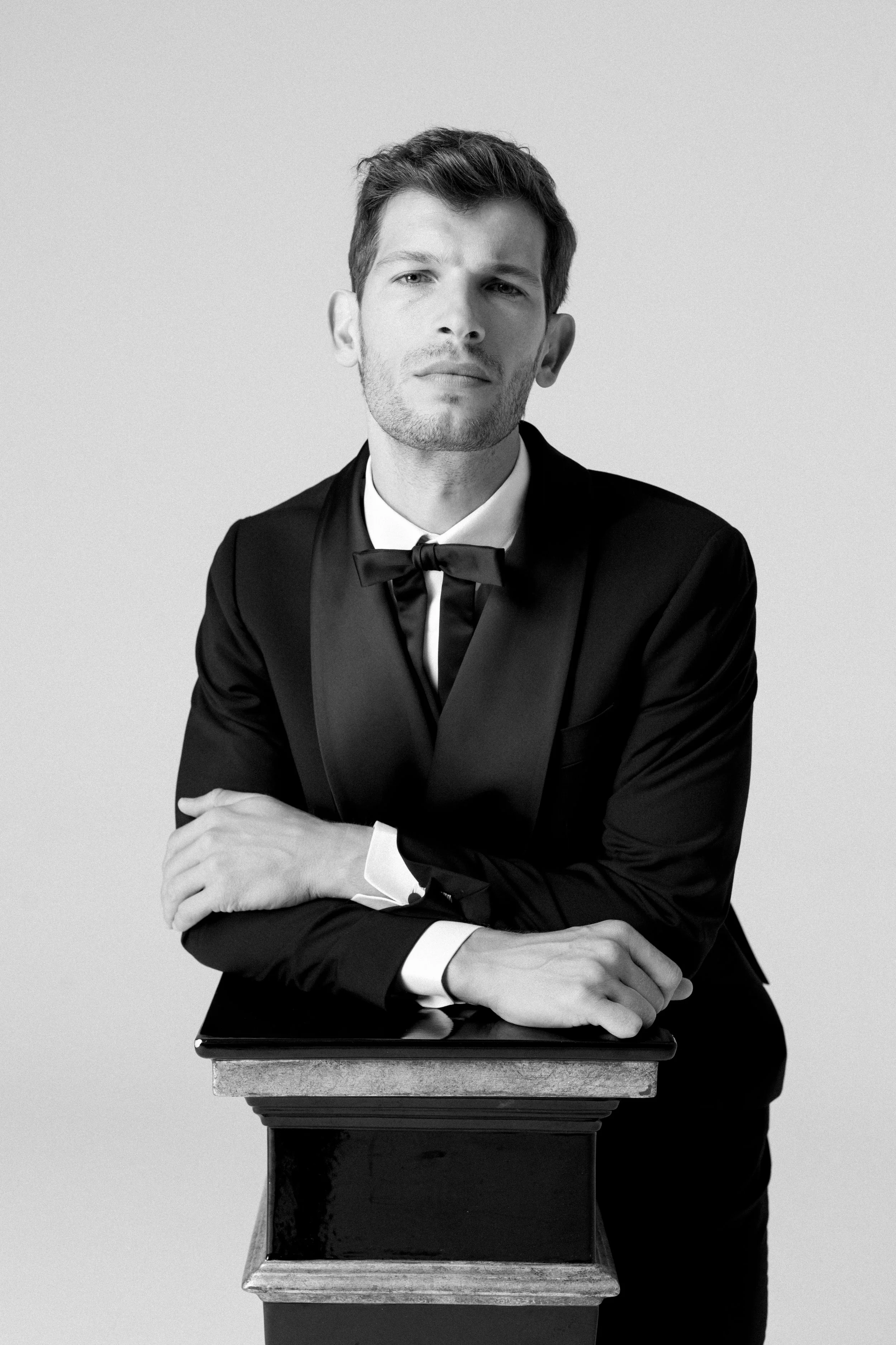 Ritratto in bianco e nero di un uomo elegante in abito con cravatta, appoggiato a un piedistallo, con espressione seria.