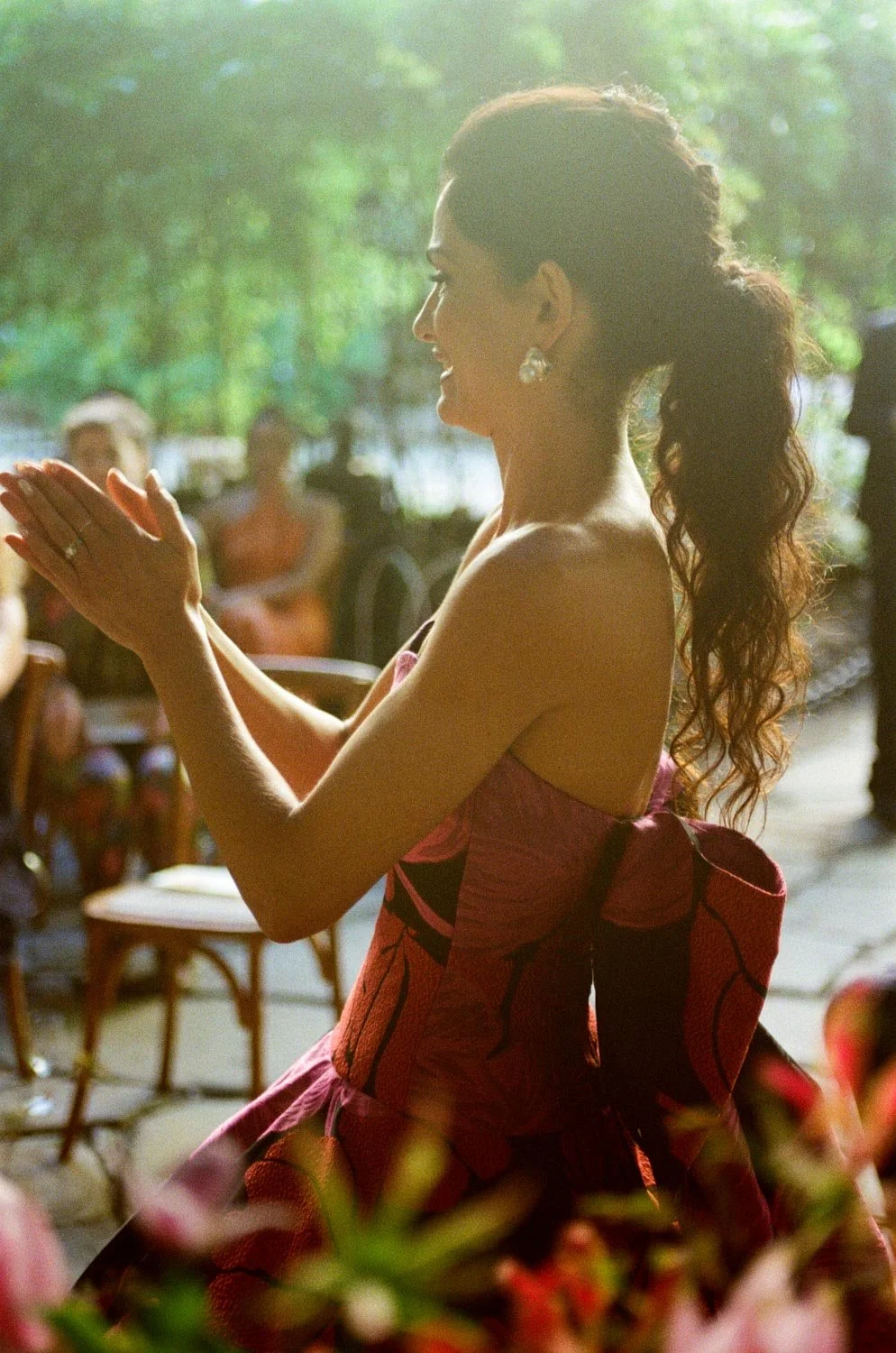 Una donna sorride e si applaude, vestita con un abito colorato. Sullo sfondo, ci sono alcune persone sedute e si intravede un ambiente naturale con alberi verdi.