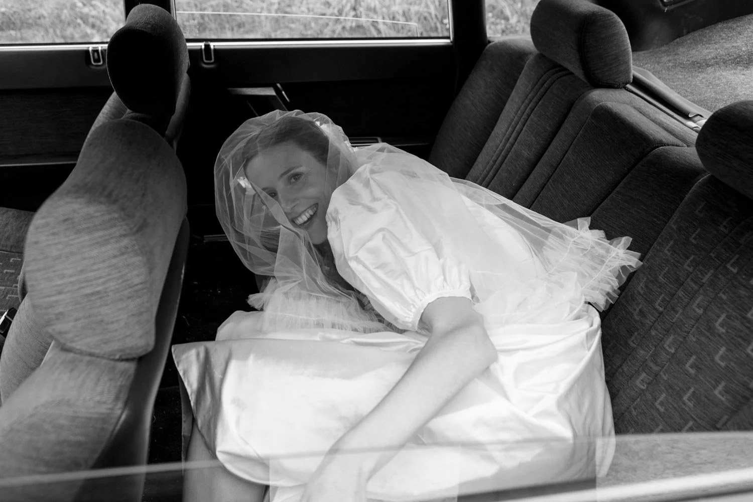 Una donna sorridente vestita da sposa si trova seduta sul sedile di un'auto, con un velo da sposa sulla testa.