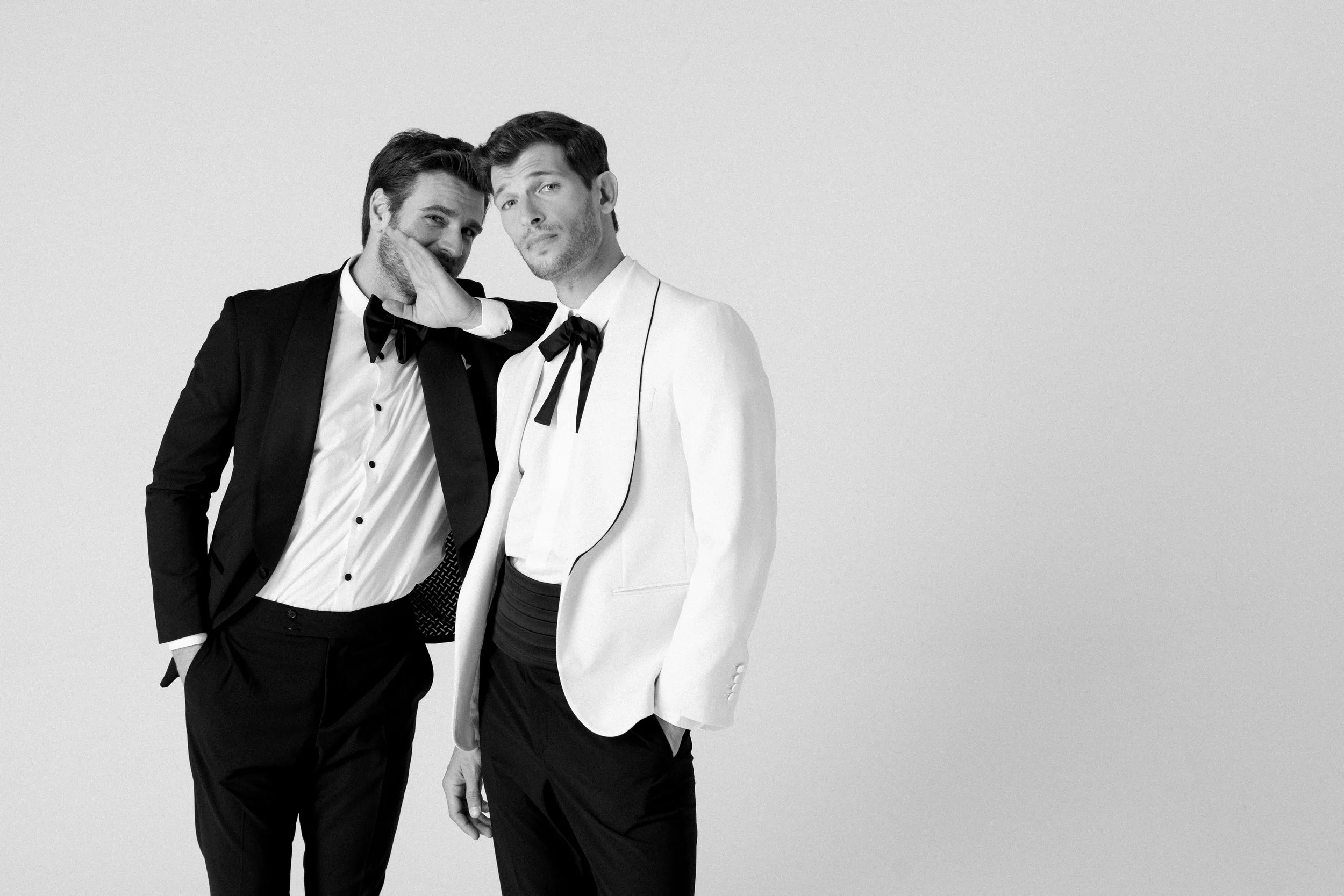 Due uomini vestiti elegantemente, uno con smoking nero e il other con smoking bianco, si trovano contro uno sfondo neutro, uno di loro fa un gesto verso l'altra con la mano aperta in modo giocoso.