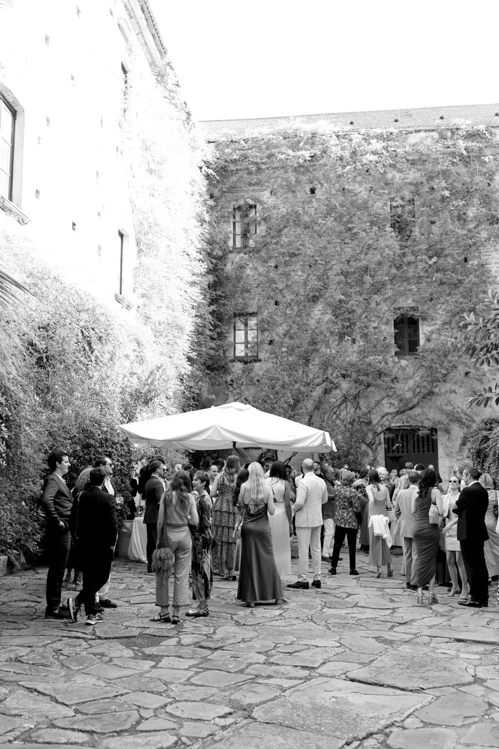 Un gruppo di persone si riuniscono in un cortile con pavimentazione di pietra, sotto un ombrellone, in un ambiente con muri ricoperti di edera e piante, forse durante un evento sociale o un ricevimento.