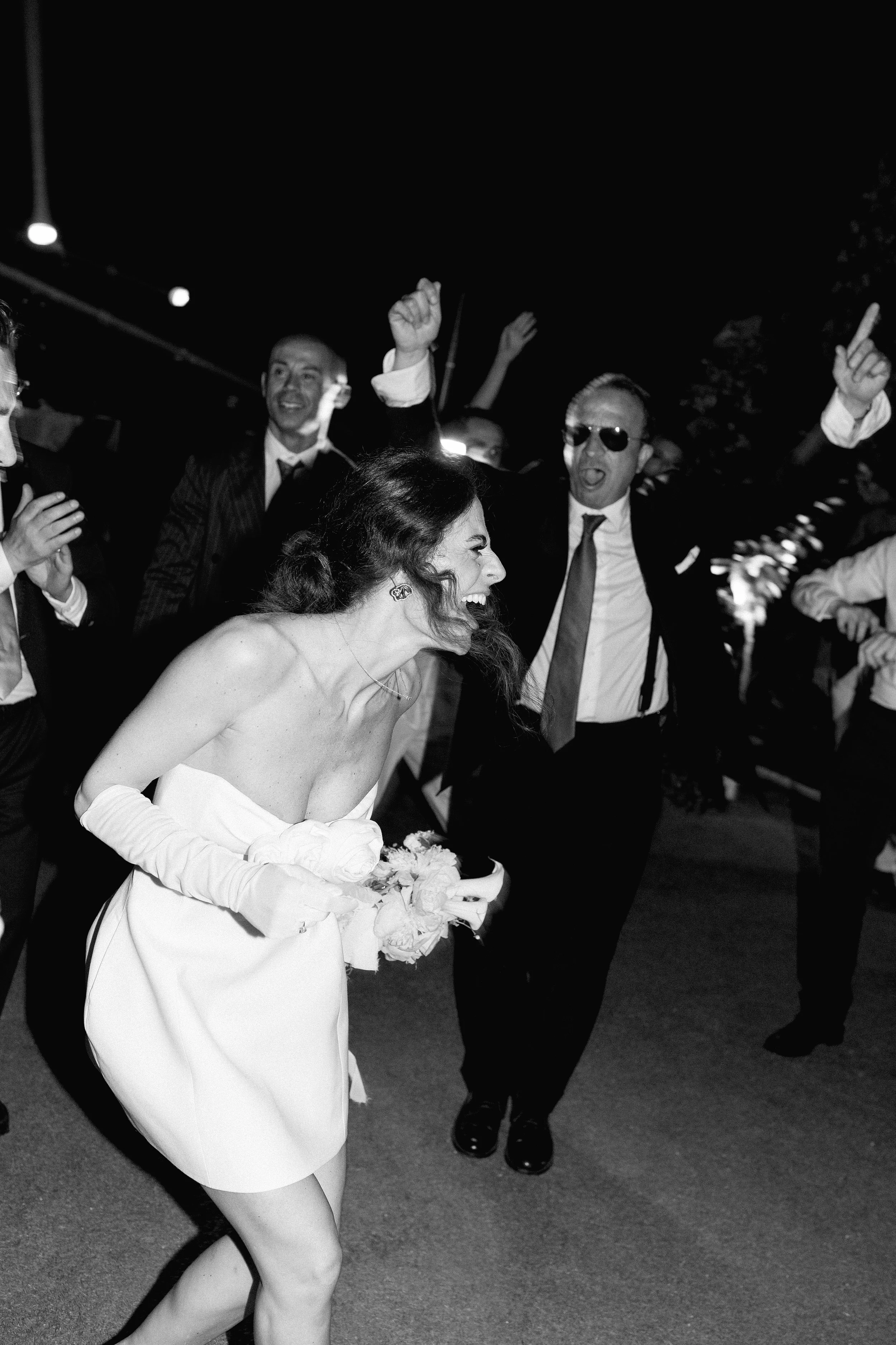 Una sposa con un abito senza spalline ride e scherza con amici durante una celebrazione notturna all'aperto.