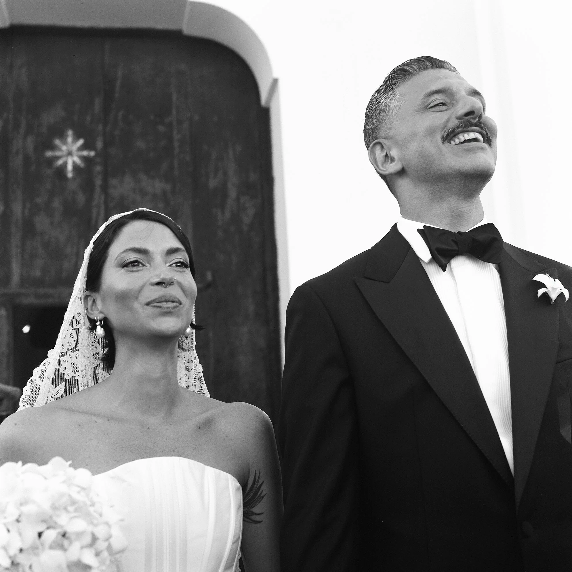 Una coppia di sposi durante il matrimonio, con la sposa che tiene un bouquet di fiori e entrambi sorridenti, in un ambiente interno.