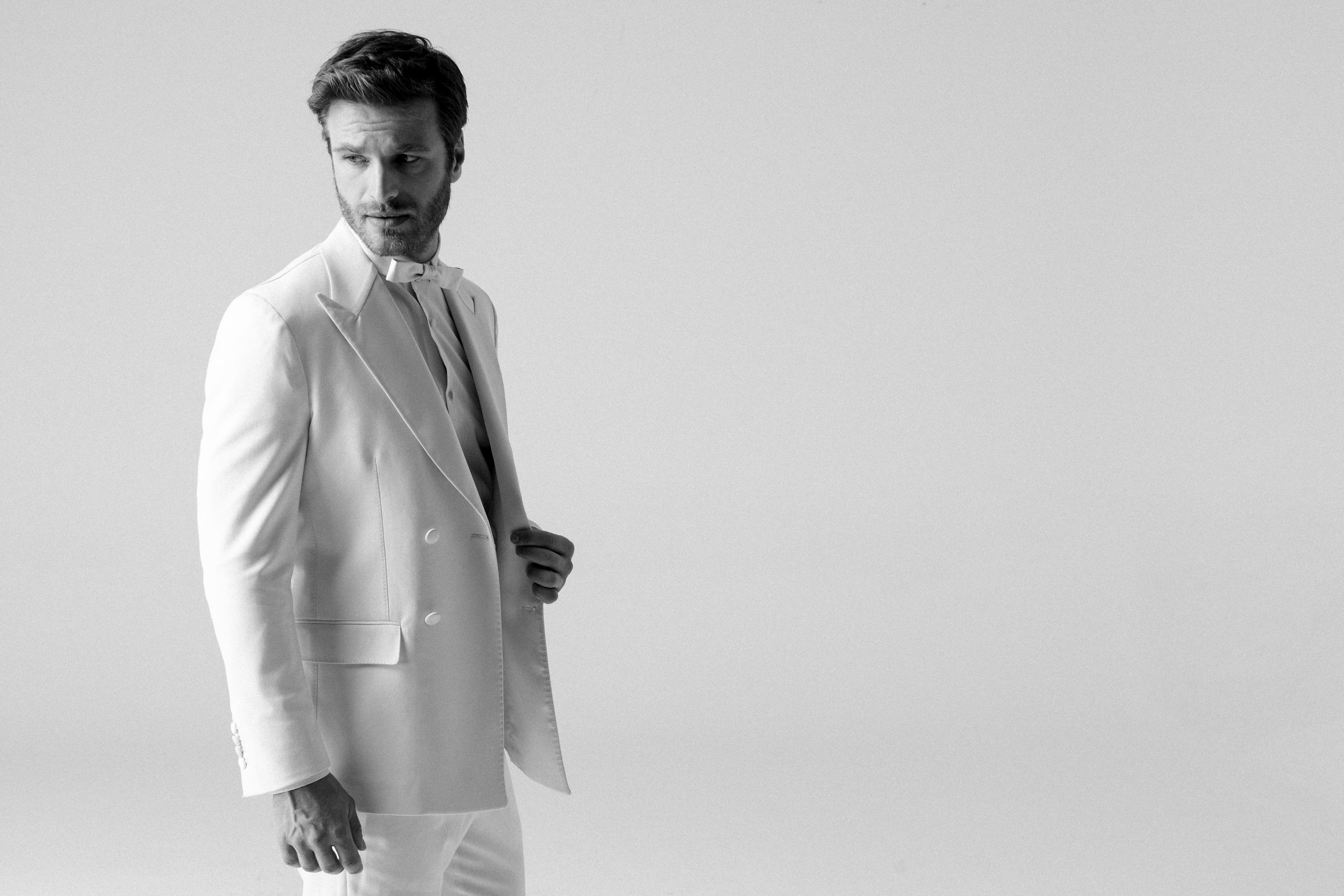 Un uomo vestito con un completo bianco, che guarda leggermente a sinistra, con sfondo neutro.