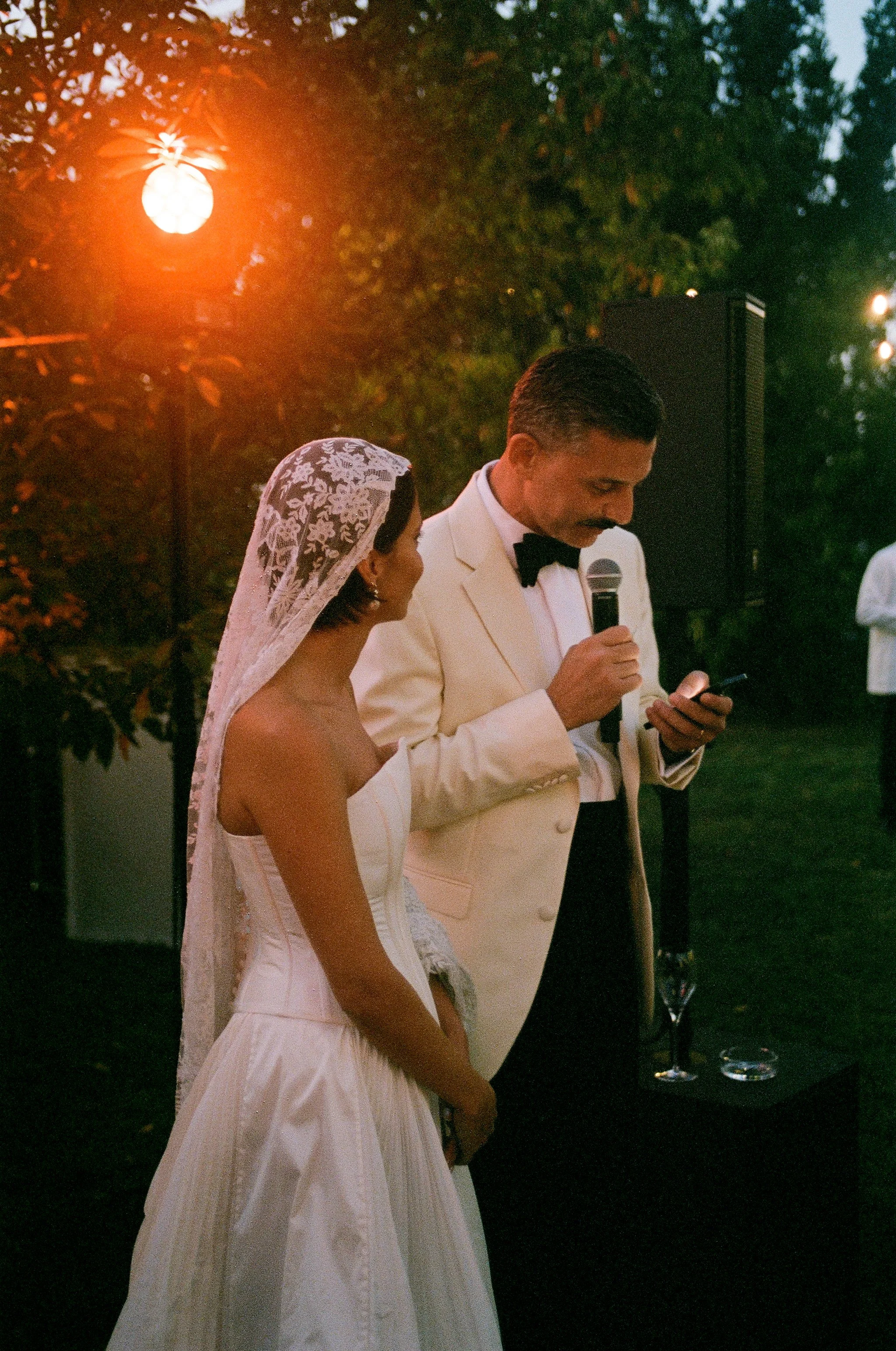 Una coppia di sposi durante il matrimonio in giardino, con un uomo vestito in tuxedo bianco e la sposa in abito bianco e mantiglia di pizzo, che ascolta mentre l'uomo legge o pronuncia un discorso.