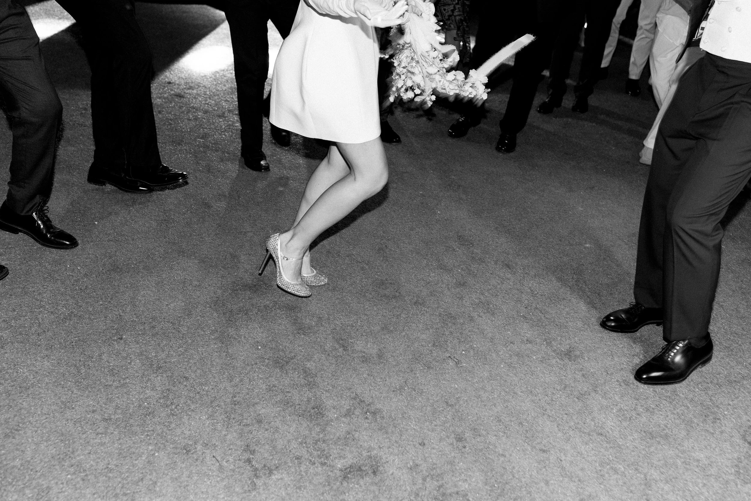 Foto in bianco e nero di persone che ballano in un ballo tradizionale, con una donna che indossa tacchi alti brillante e un vestito chiaro.