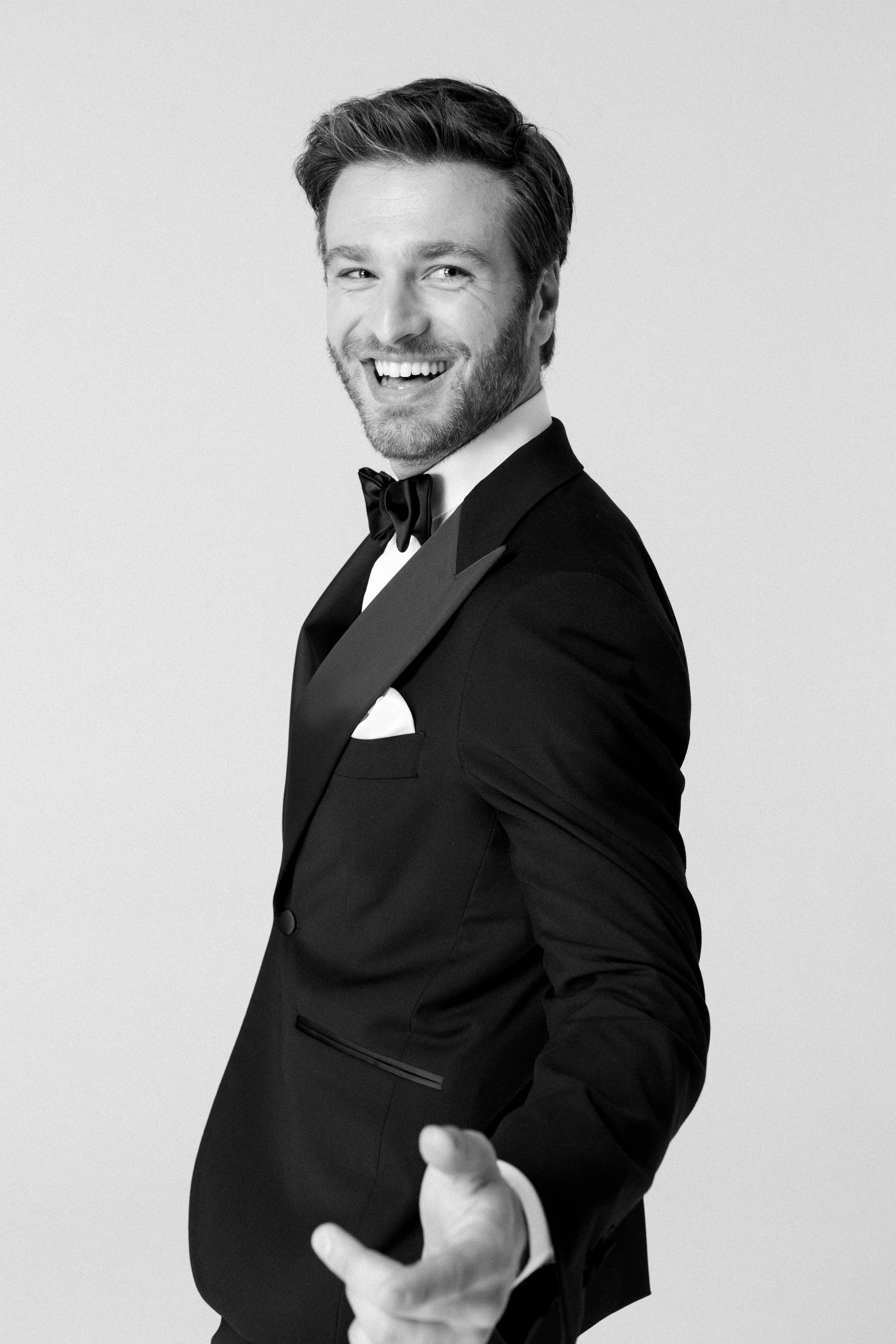 Uomo con abito da sera nero e papillon che sorride e indica verso l'obiettivo.