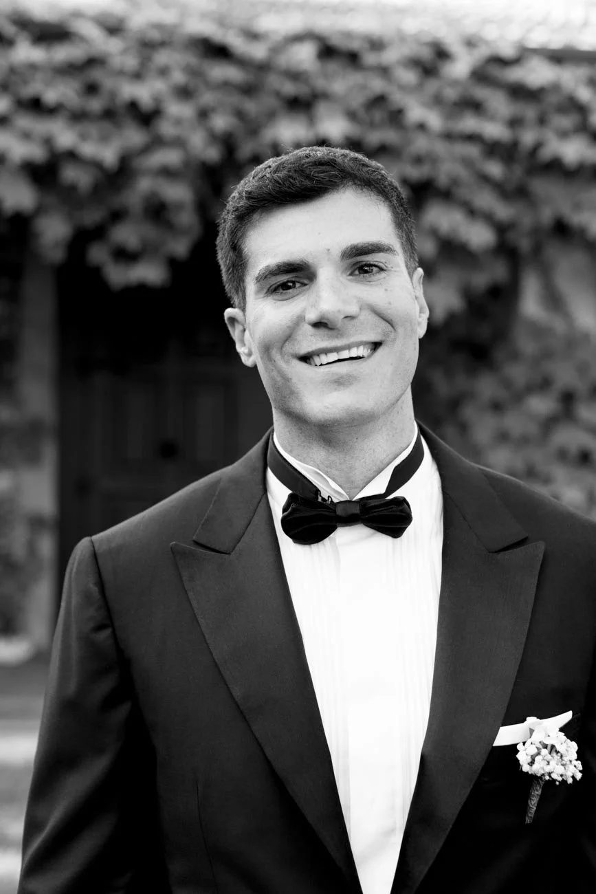 Un uomo sorridente in completo da sera con papillon nero, abbigliamento da matrimonio, in un ambiente esterno con facciata di un edificio di pietra in background.