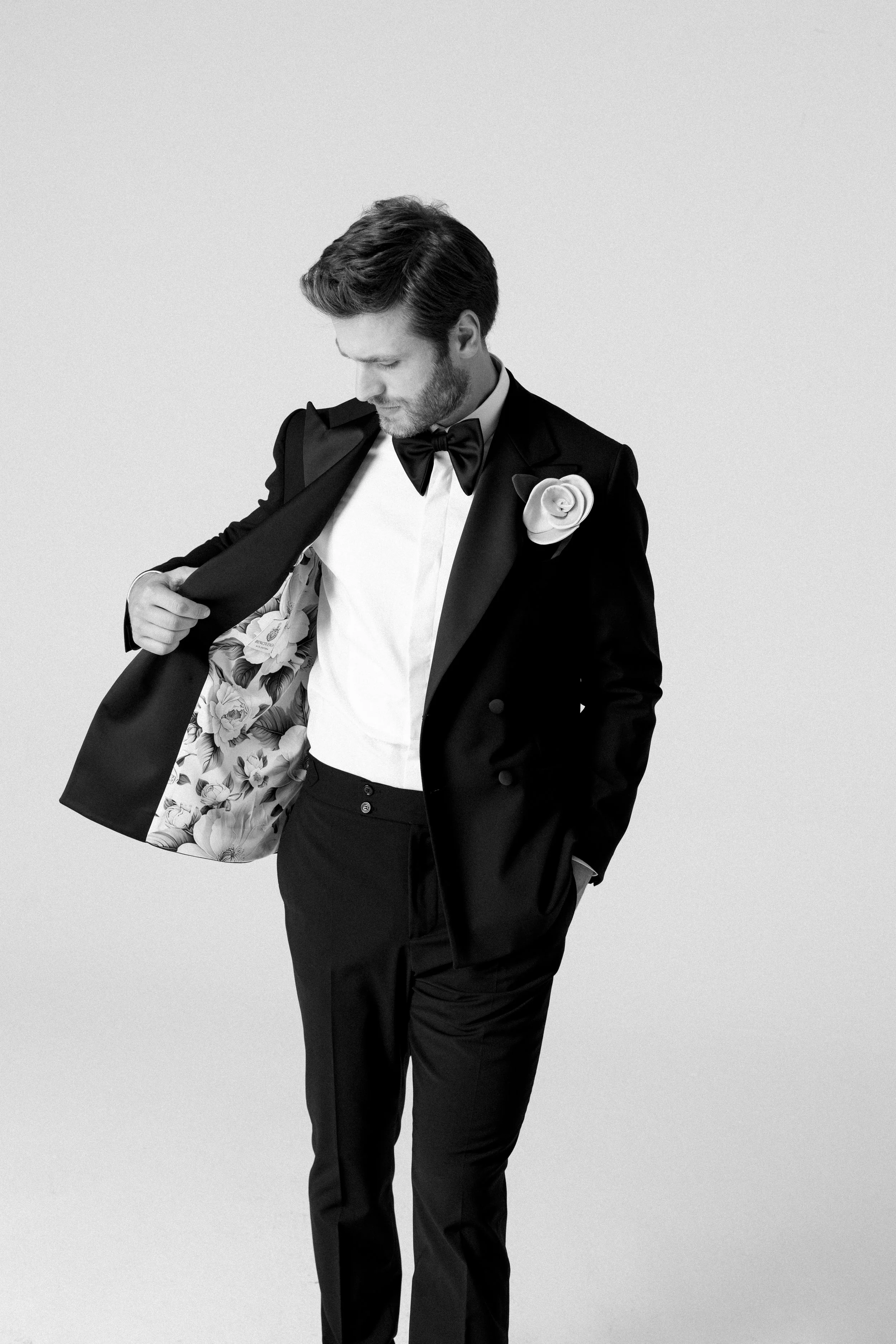 Uomo in abito da sera nero con papillon nero e fiore sulla giacca, che apre la giacca mostrando una fodera con stampa floreale, sfondo bianco.