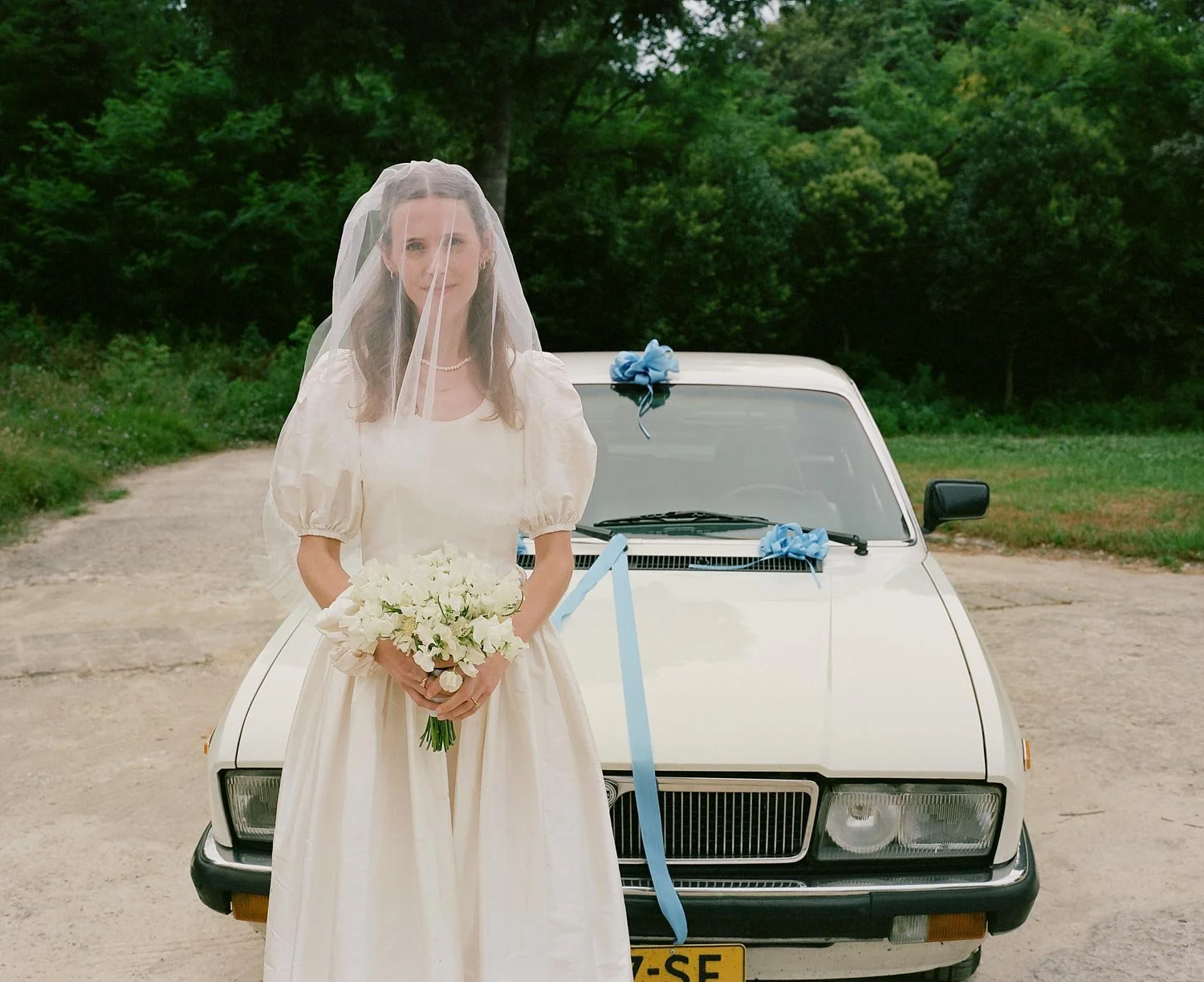Una sposa con un abito bianco, velo e bouquet di fiori, davanti a un'auto decorata con nastri blu, su una strada di campagna con alberi sullo sfondo.