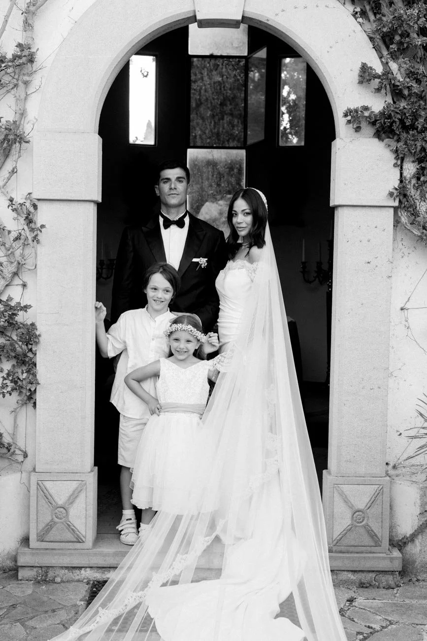 Una coppia di sposi e due bambine sorridenti, immersi in una cornice di un archivio di matrimonio, con l'architettura in pietra e viti che crescono sulla parete.