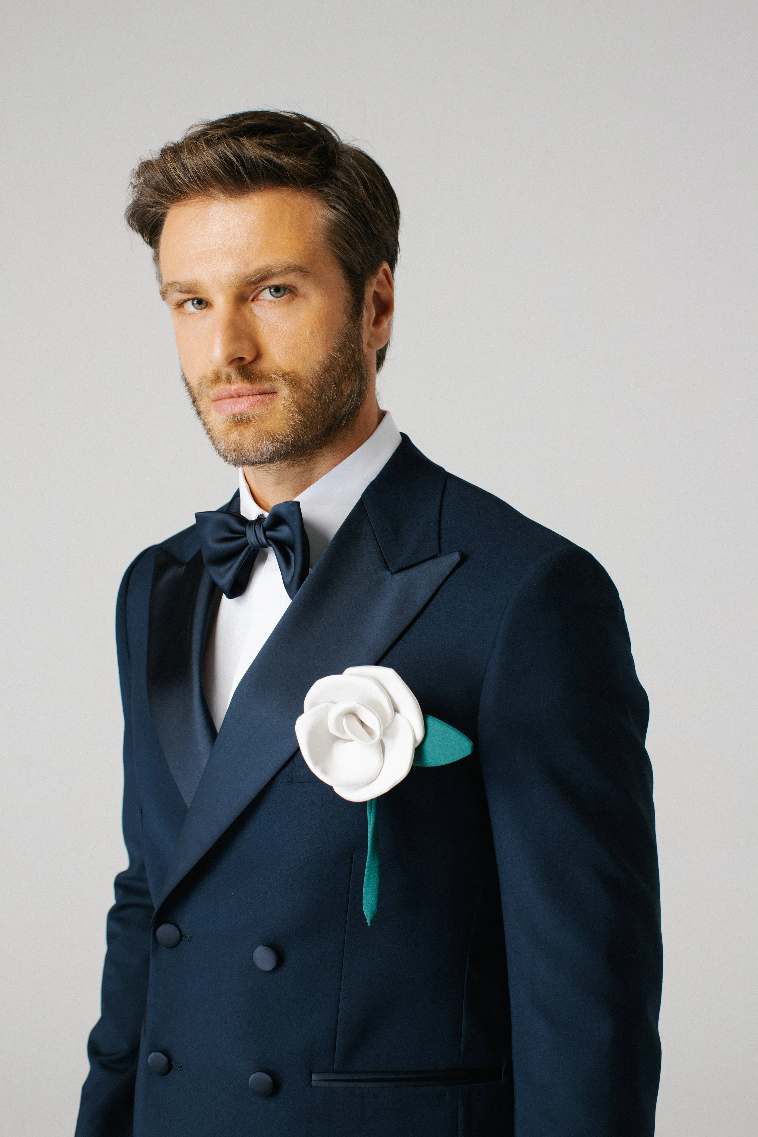 Un uomo con abito elegante blu scuro, cravatta e fiocco, con un fiore bianco sulla giacca.