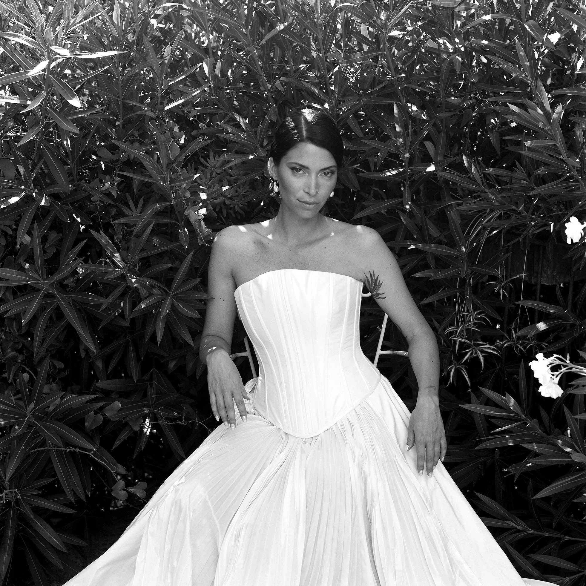 Una donna in abito bianco da sera seduta davanti a un cespuglio folto di foglie.