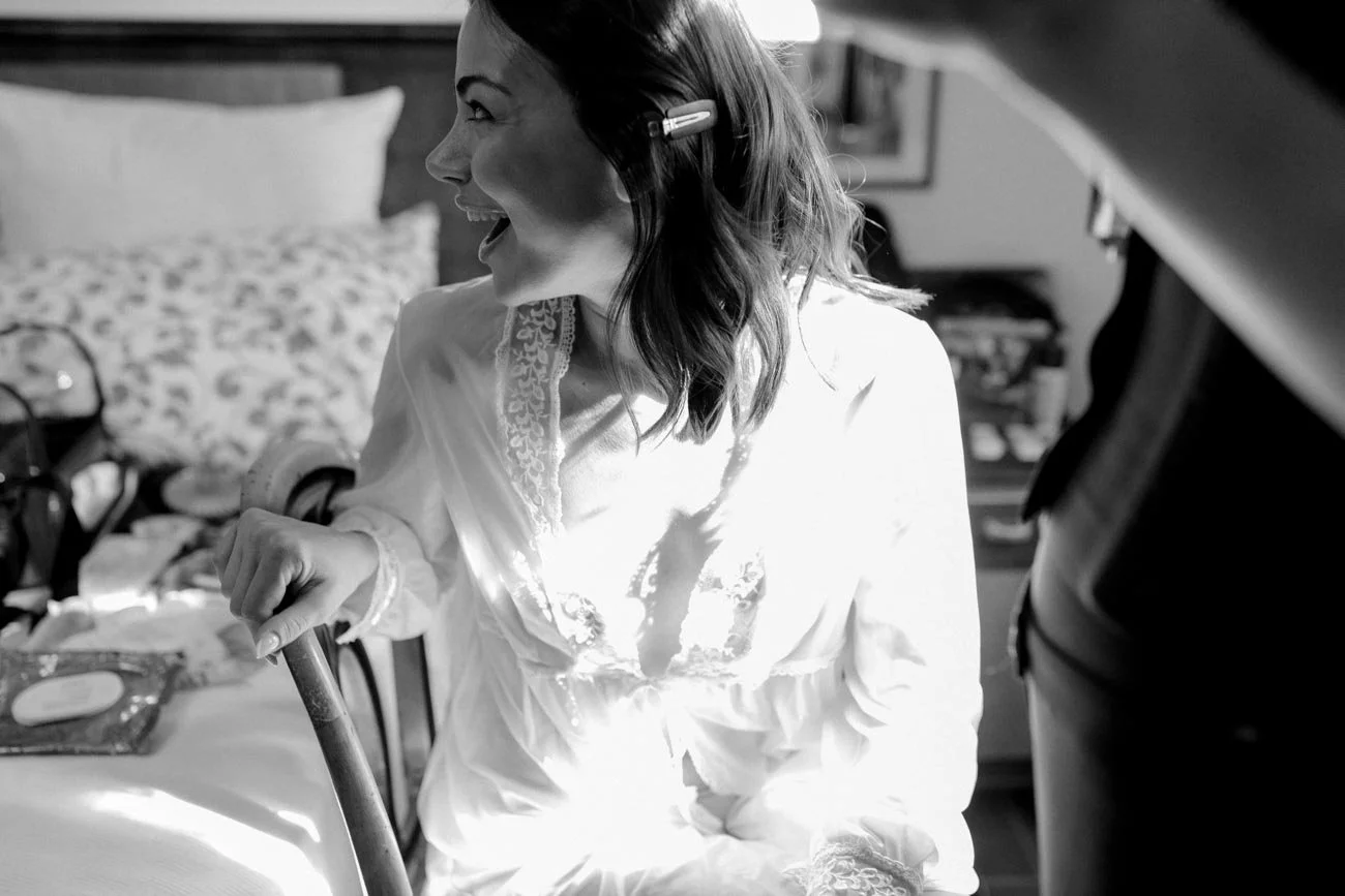 Ragazza che ride, seduta a un tavolo, con capelli castani mossi, vestita con una camicia bianca con dettagli in pizzo, in una stanza con un letto sullo sfondo.