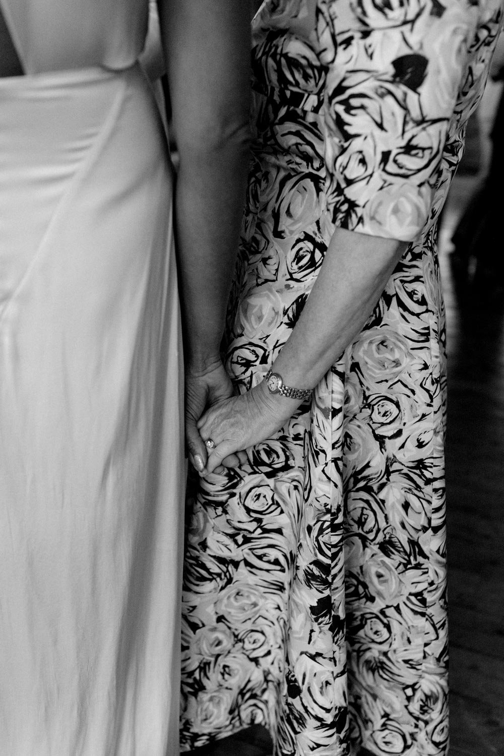 Due persone si tengono per mano, una vestita con un abito bianco e l'altra con un completo a fantasia con fiori.