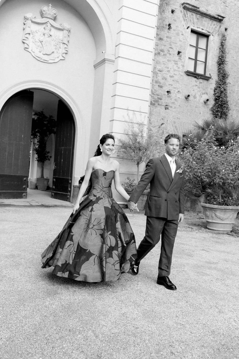 Una coppia vestita in abiti eleganti cammina insieme fuori da un edificio storico, con piante sullo sfondo.