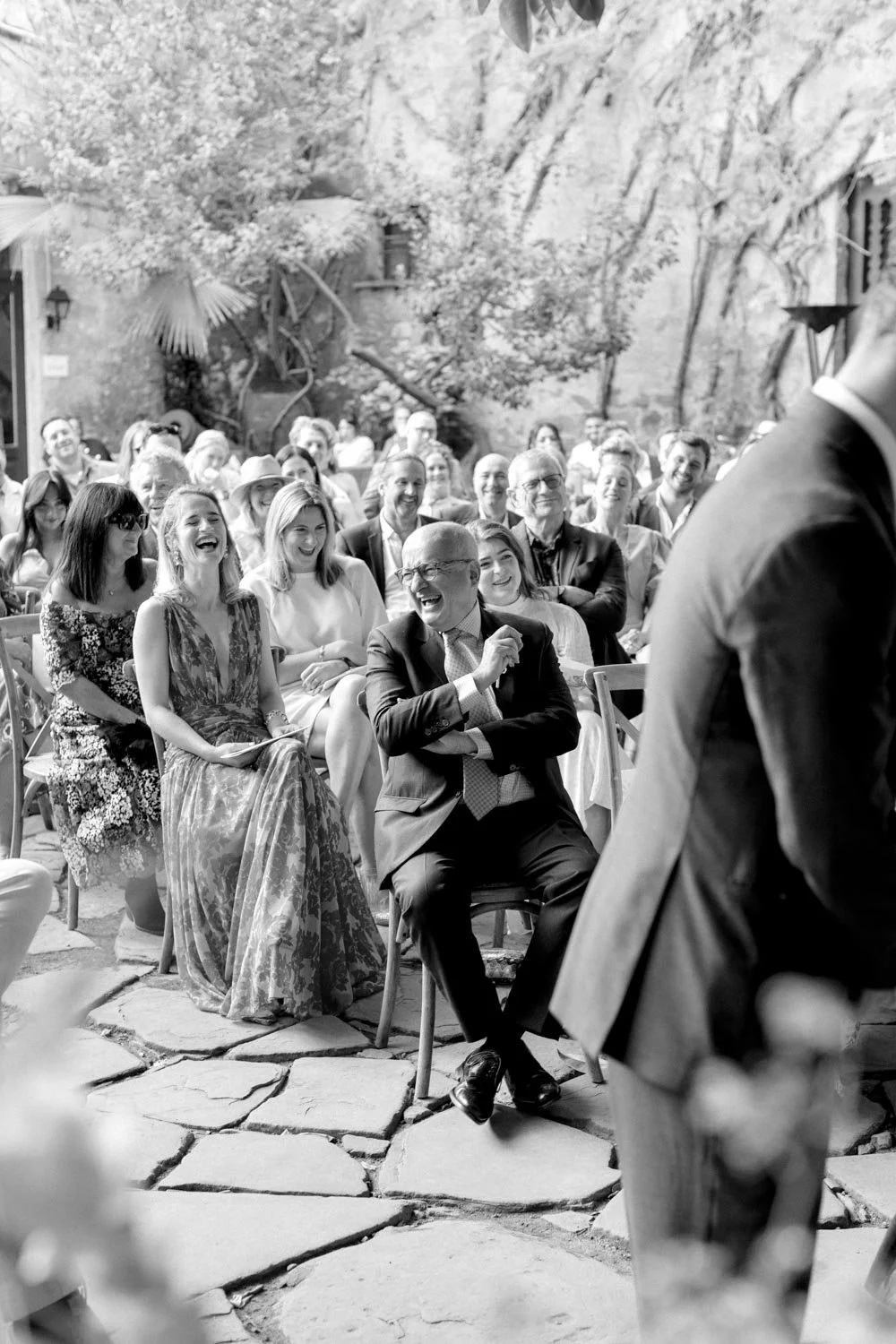 Foto in bianco e nero di un matrimonio all'aperto con ospiti seduti che ridono e sorridono, mentre la coppia protagonista si trova di fronte.