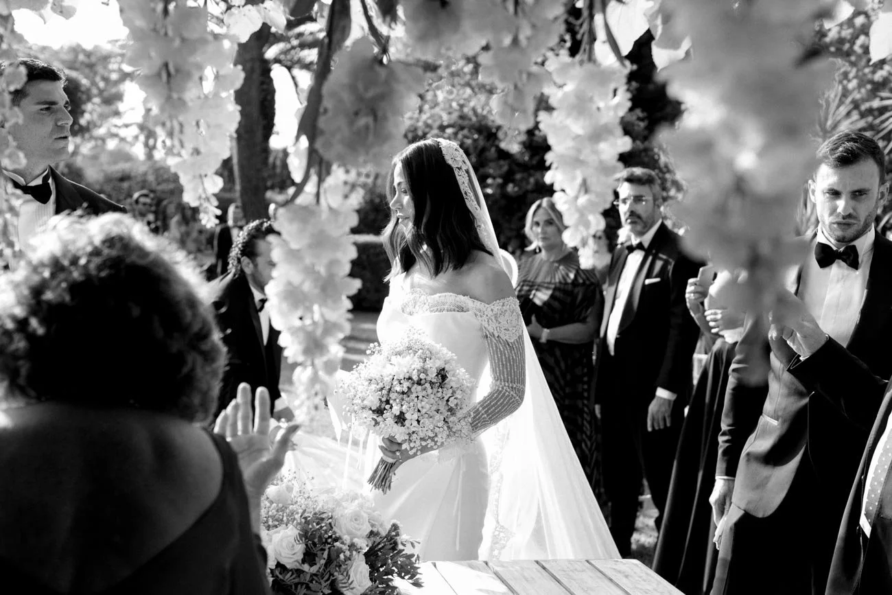 Una coppia di sposi al matrimonio immersi in un contesto all'aperto, circondati da altri invitati. La sposa indossa un abito bianco con velo e tiene un bouquet di fiori, mentre il partner e gli invitati sono vestiti elegantemente in abiti da sera.