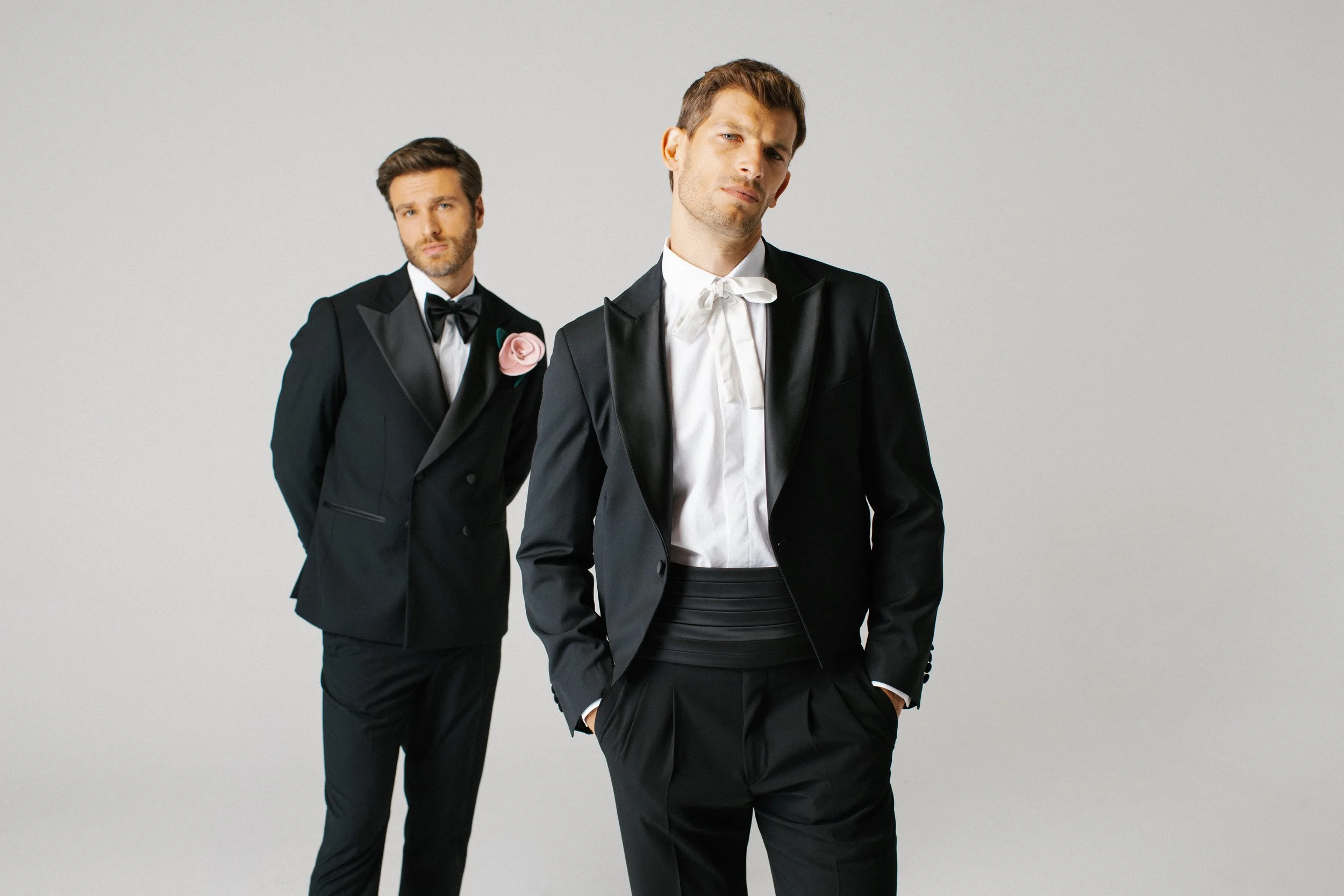 Due uomini in abiti da sera eleganti, uno in primo piano con mano in tasca e l'altro sullo sfondo con fiore rosa sulla giacca, entrambi con espressioni serie, contro uno sfondo grigio chiaro