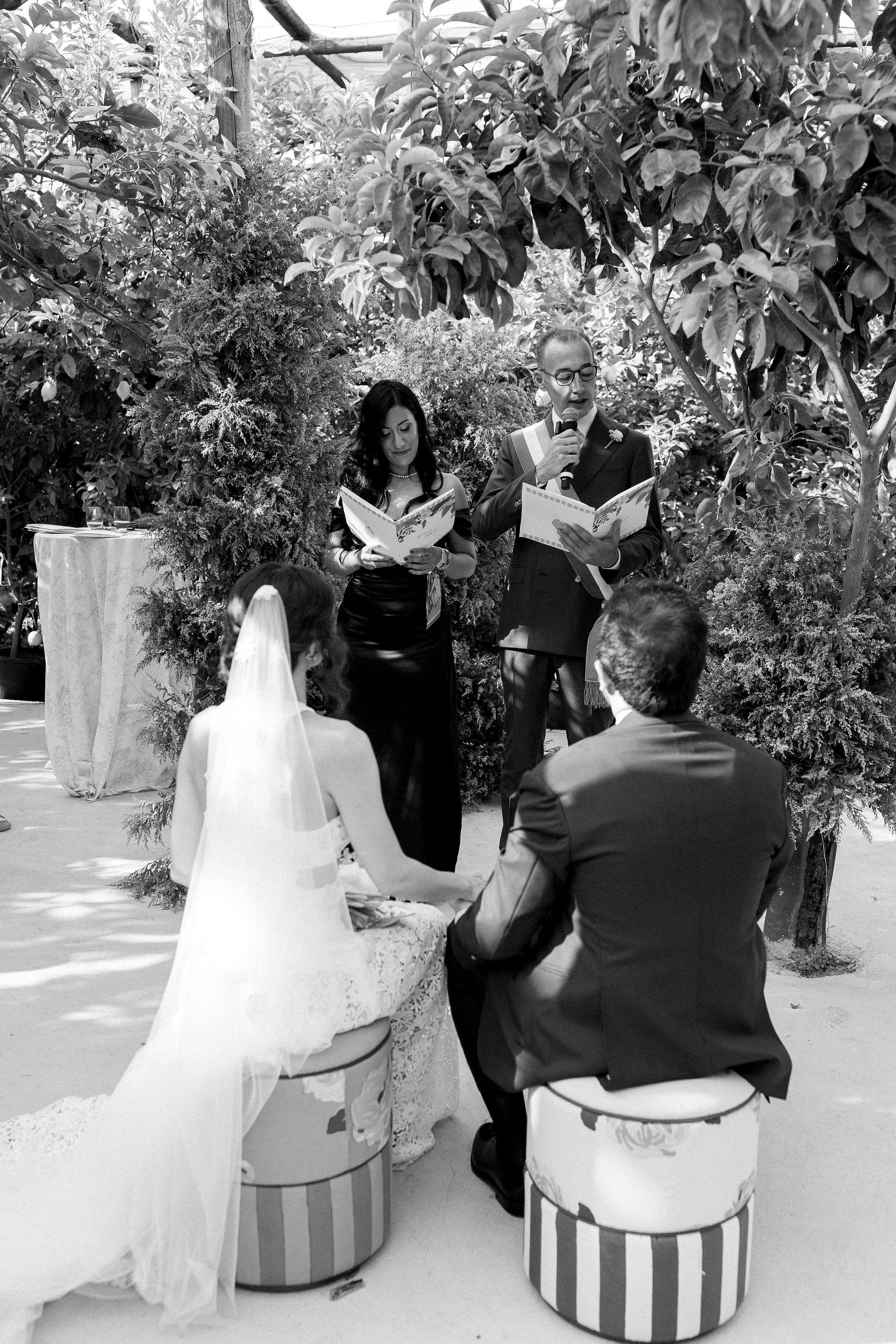 Una coppia di sposi seduta durante un matrimonio circondata da vegetazione, con due persone che leggono i voti o un testo della cerimonia, una delle quali con un microfono.
