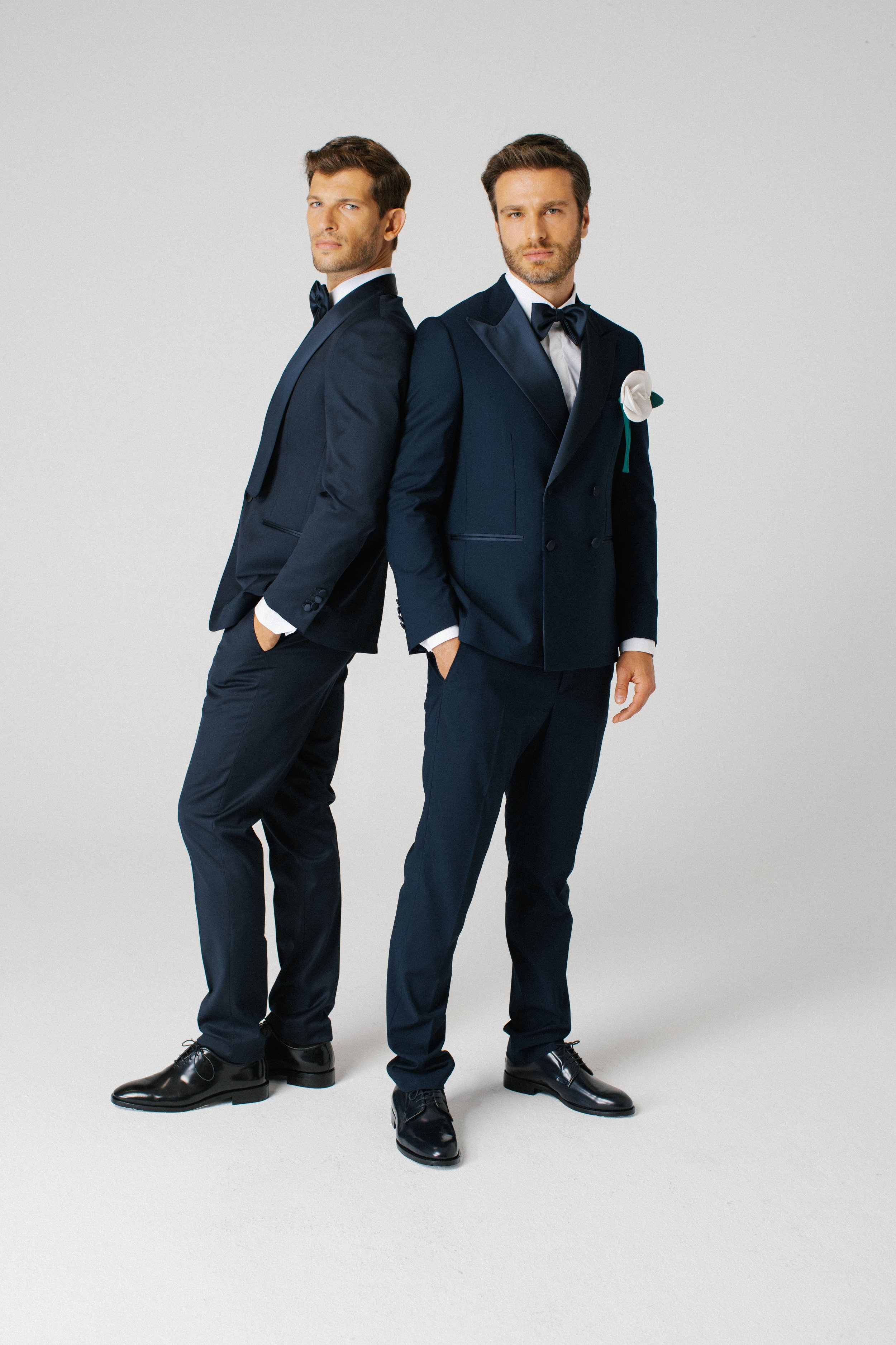 Due uomini vestiti con abiti da sera eleganti, uno con un papillon, in piedi uno davanti all'altro in un ambiente minimale e senza sfondo.