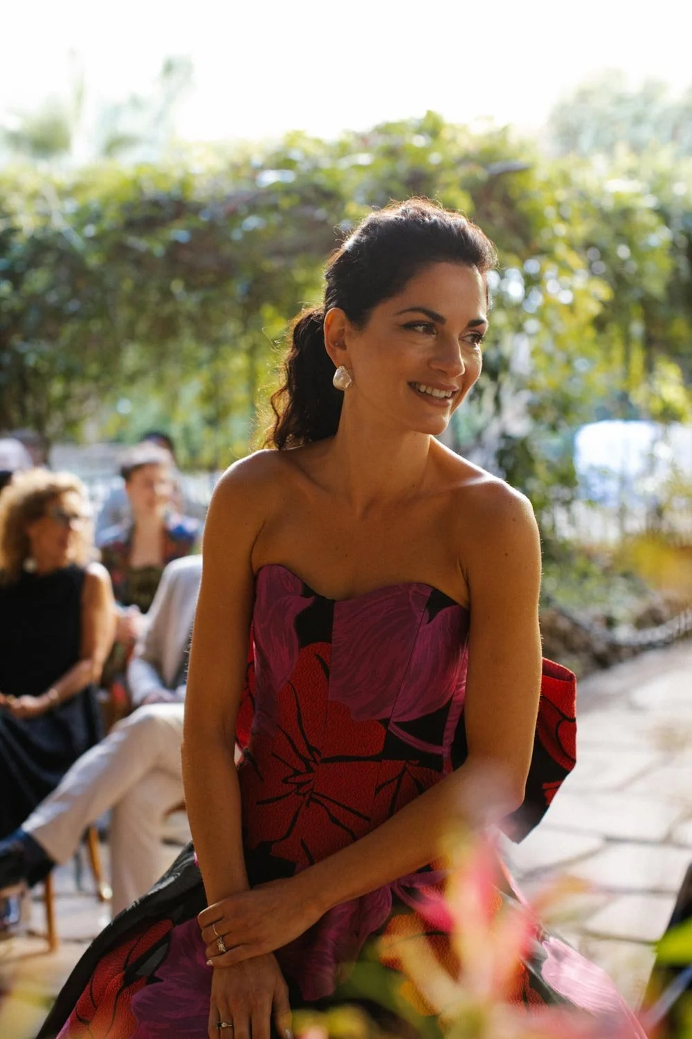 Una donna sorridente con capelli scuri e leggermente ricci vestita con un abito rosso con motivi floreali, seduta tra altre persone in un ambiente all'aperto con alberi e luce naturale.