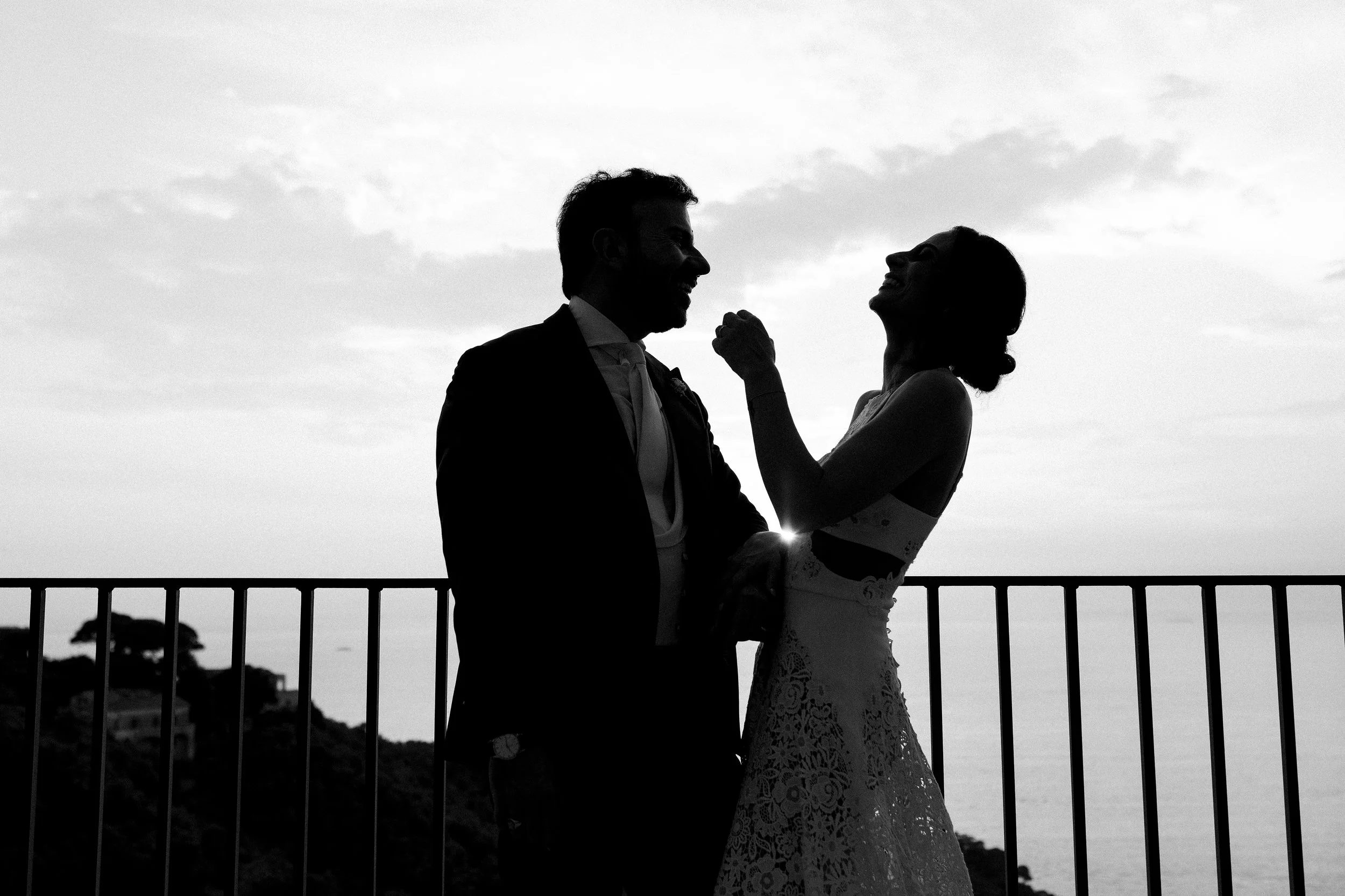 Silhouette di una coppia in abiti da matrimonio che ride e si tiene per mano durante il tramonto, con uno sfondo di cielo nuvoloso