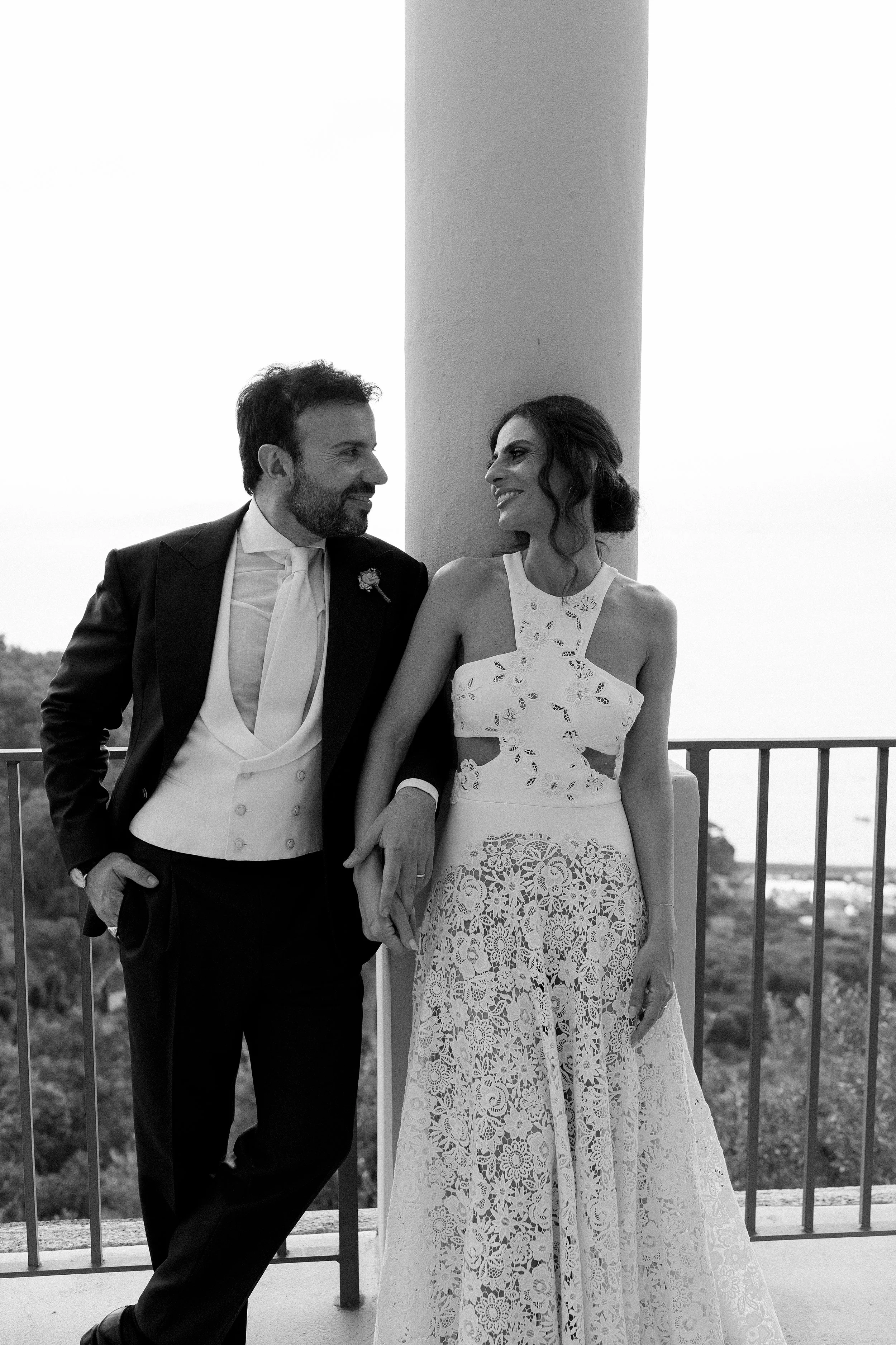 Una coppia di sposi si guarda sorridendo, mano nella mano, in un'ambientazione esterna, con un pilastro e un panorama sullo sfondo.