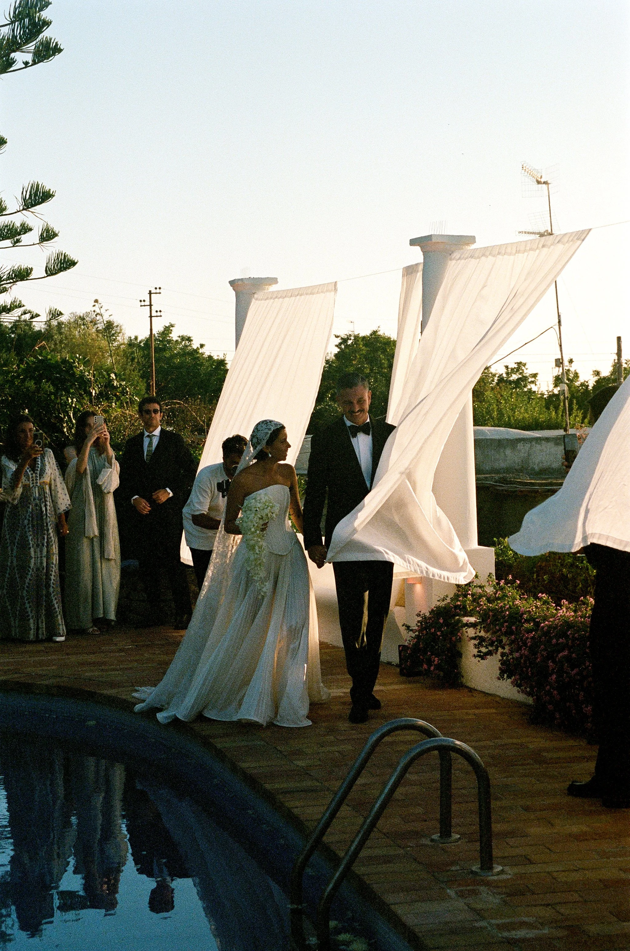 Una coppia di sposi sorride tenendosi per mano durante il matrimonio, circondati da amici e parenti, all'aperto vicino a una piscina, con decorazioni e piante.