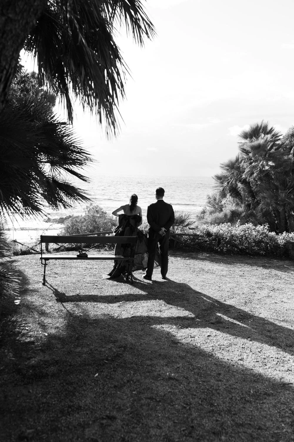 Due persone in abiti eleganti vicino al mare, con alberi e vegetazione in primo piano, vista in lontananza dell'oceano al tramonto o all'alba.