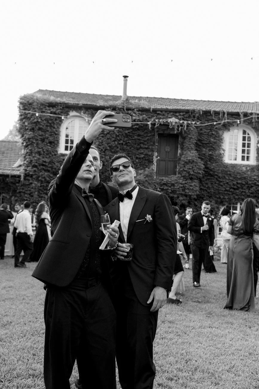 Due uomini in abiti eleganti fanno un selfie durante una festa all'aperto con molte persone sullo sfondo.