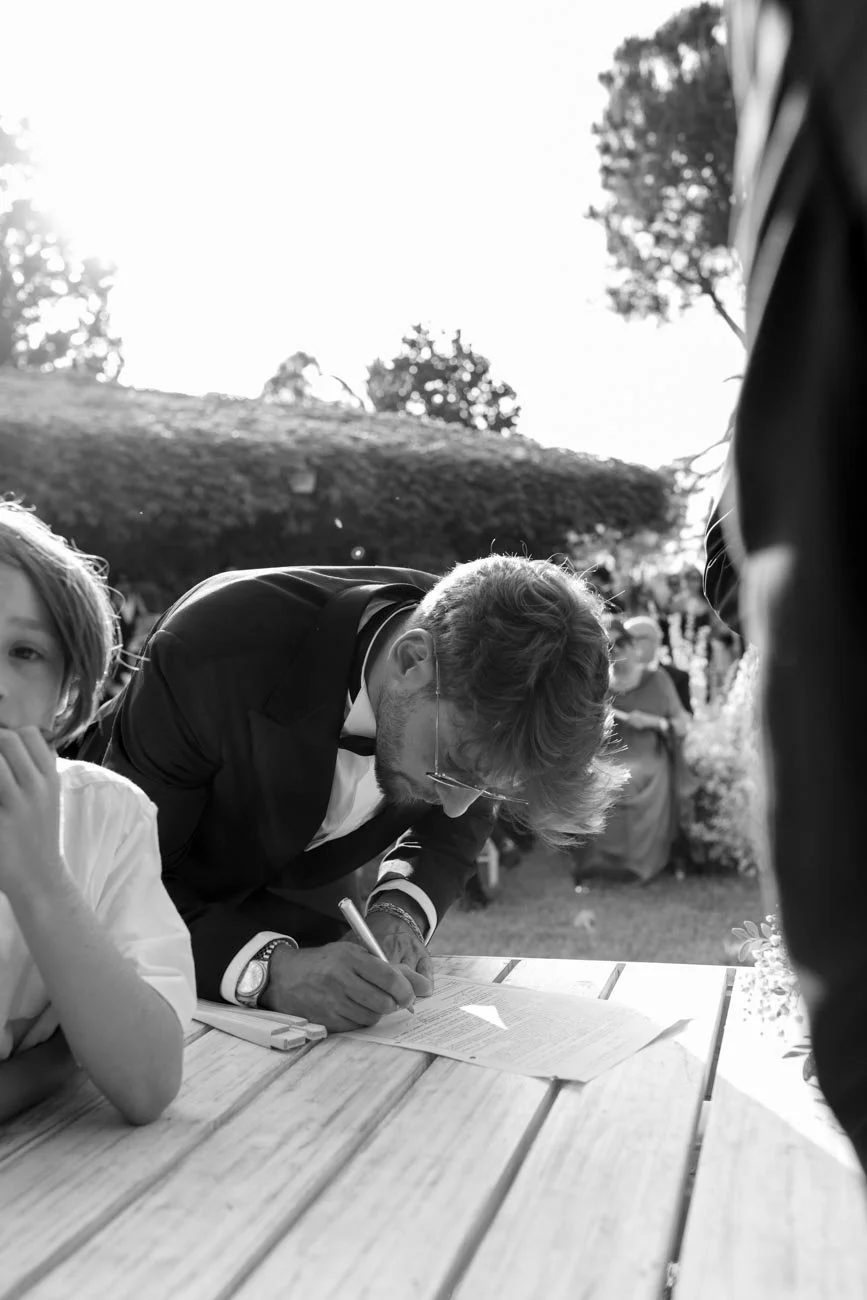 Un uomo in abito firmato firma un documento su un tavolo di legno all'aperto, con un bambino seduto accanto, durante un evento all'aperto al tramonto.