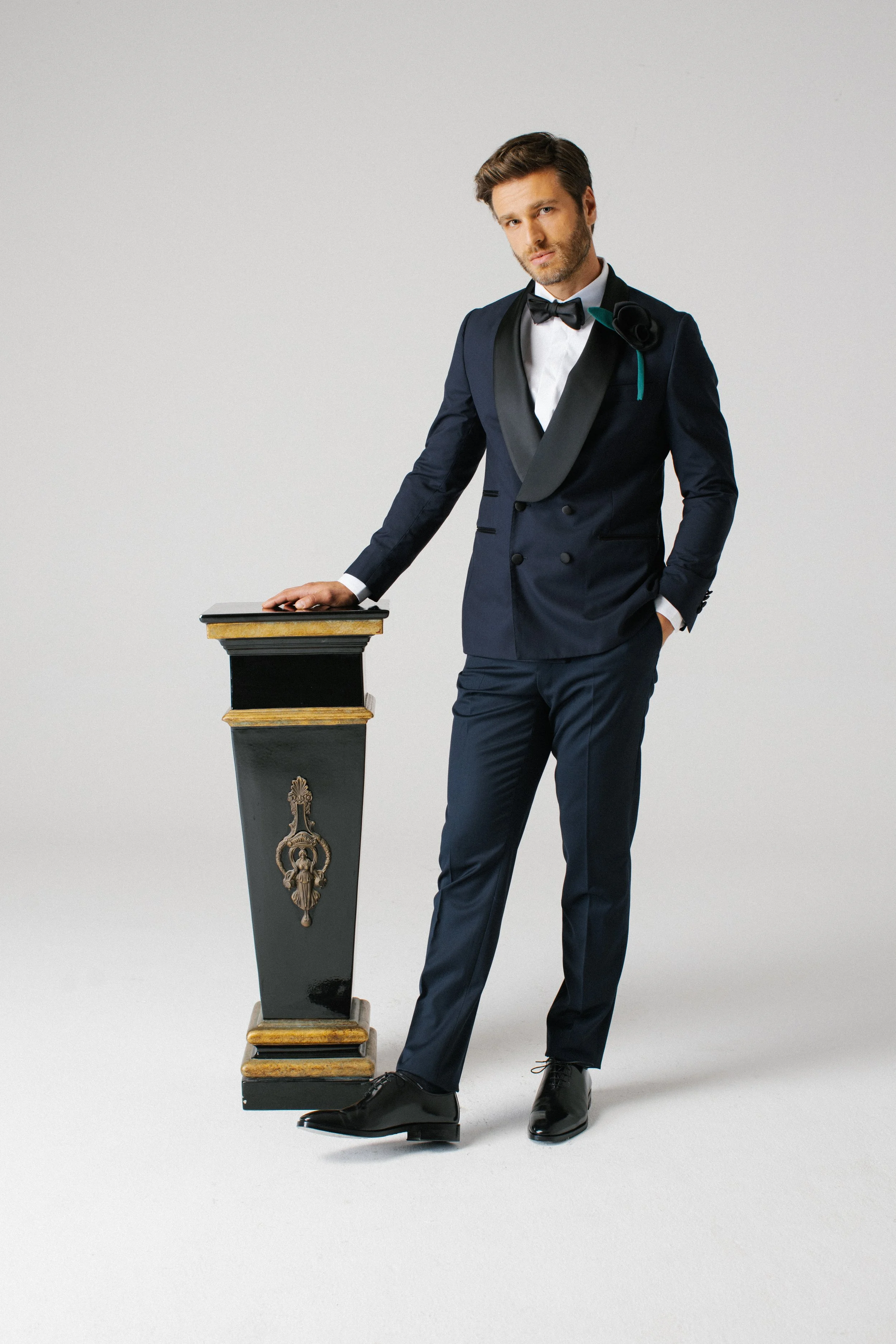 Uomo eleganti in completo da sera con papillon nero, posa con mano sulla sedia decorata, sfondo neutro