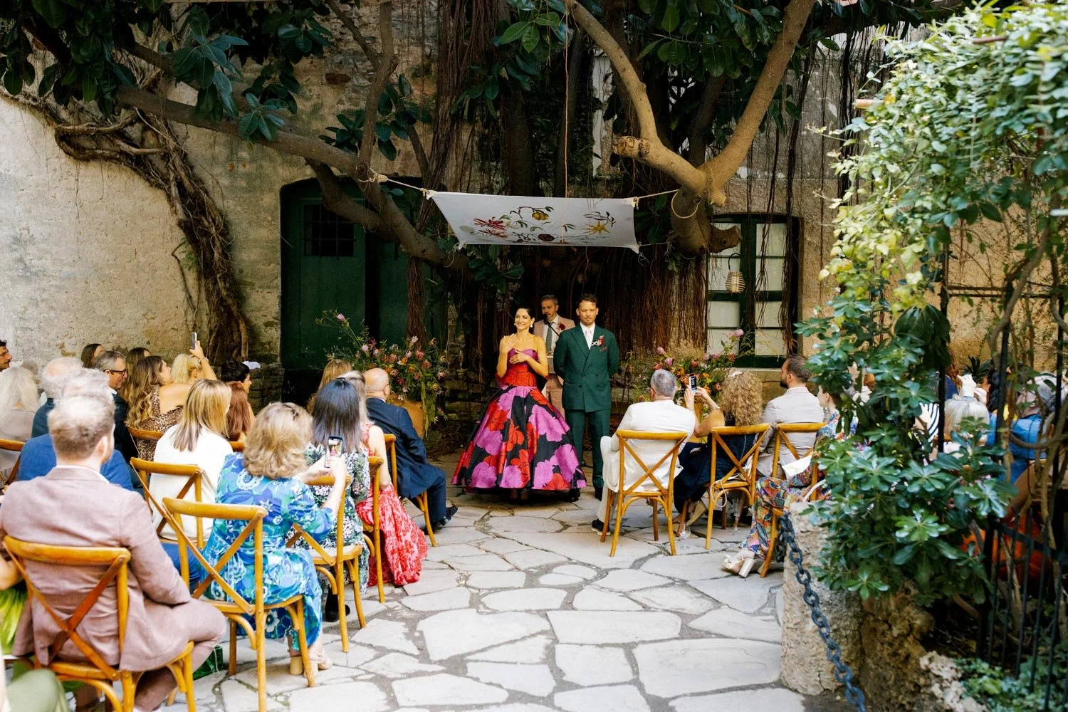 Cerimonia di matrimonio all'aperto con sposi che si tengono per mano sotto un albero, circondati da invitati seduti su sedie di legno, in un cortile con mura di pietra e vegetazione.