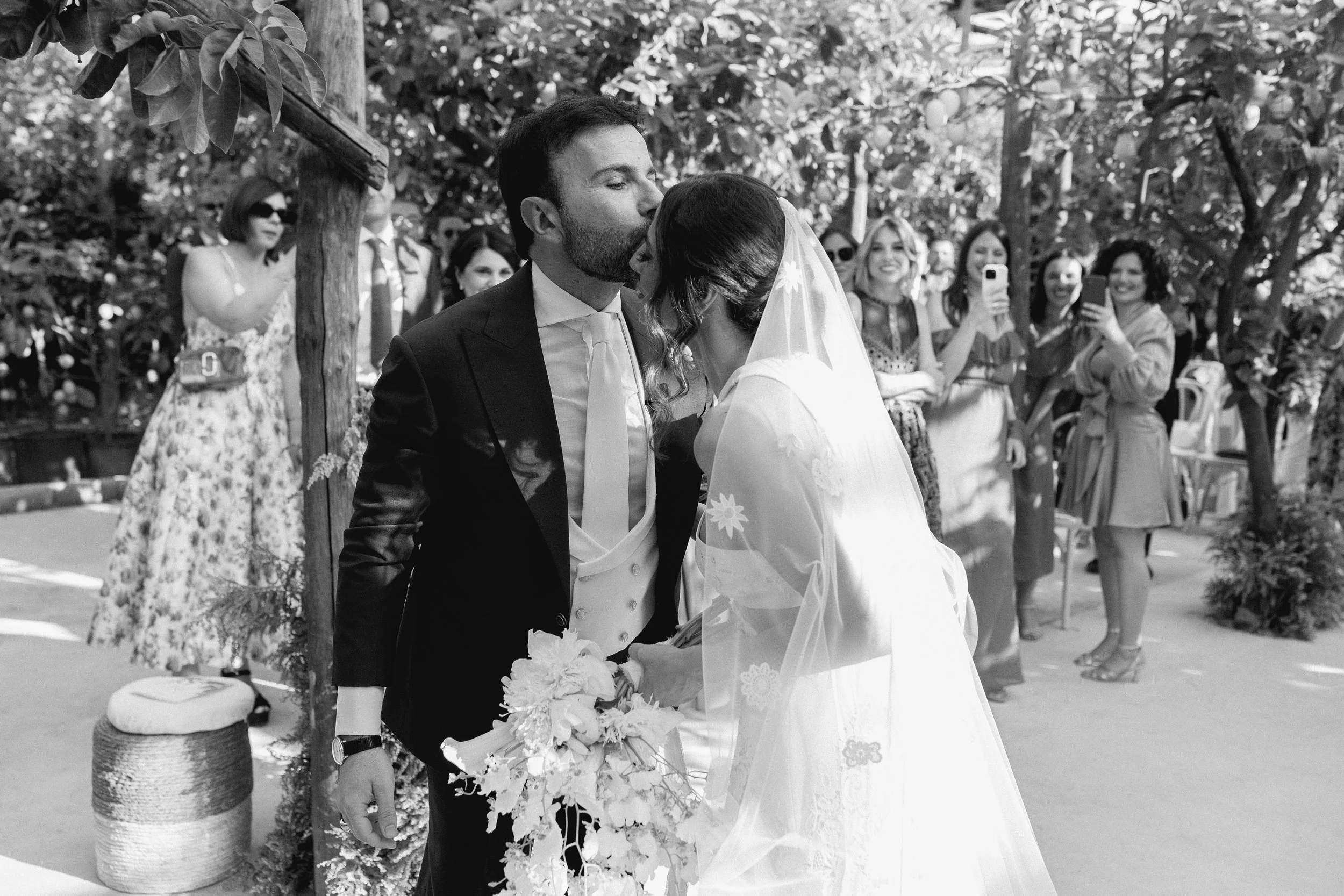 Il matrimonio in un cortile con un uomo in abito classico e una donna in abito da sposa che si baciano davanti a un gruppo di persone che scattano foto e guardano con sorrisi.